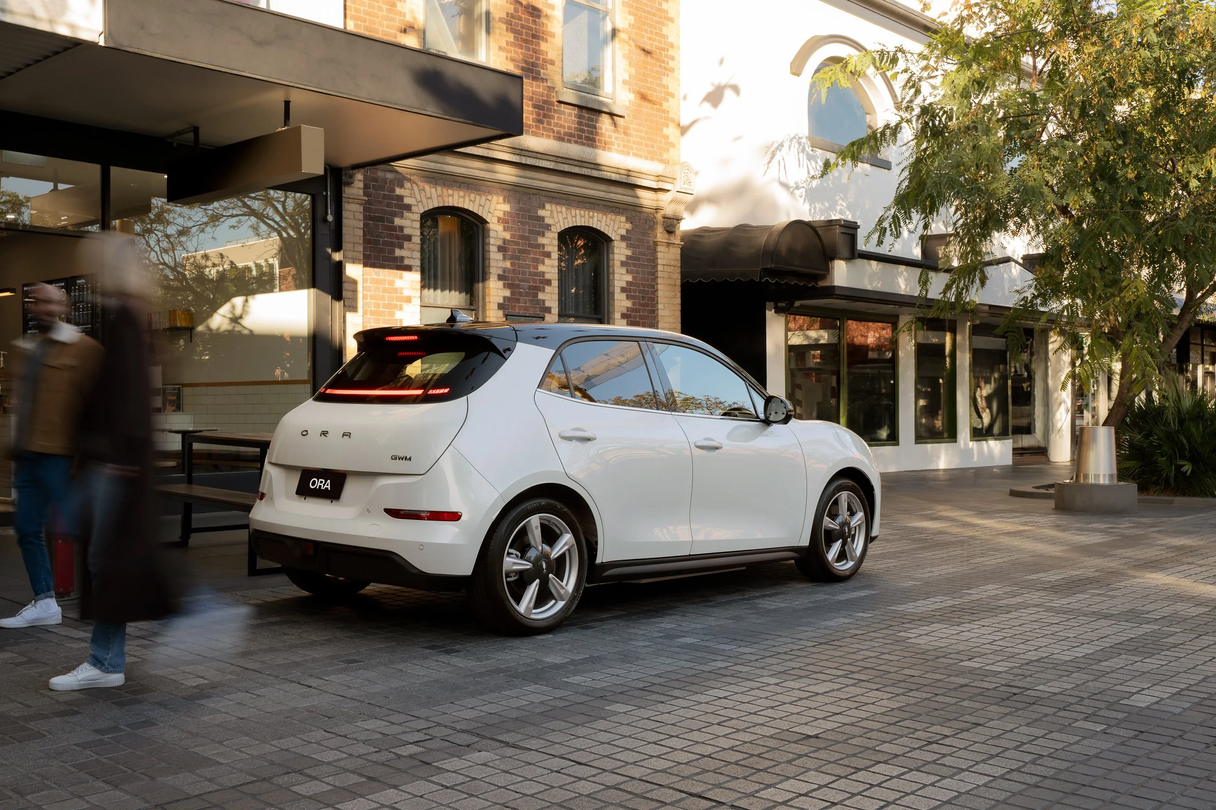 A white electric car parked on a city street in front of a brick building with shop windows, trees, and people walking by.