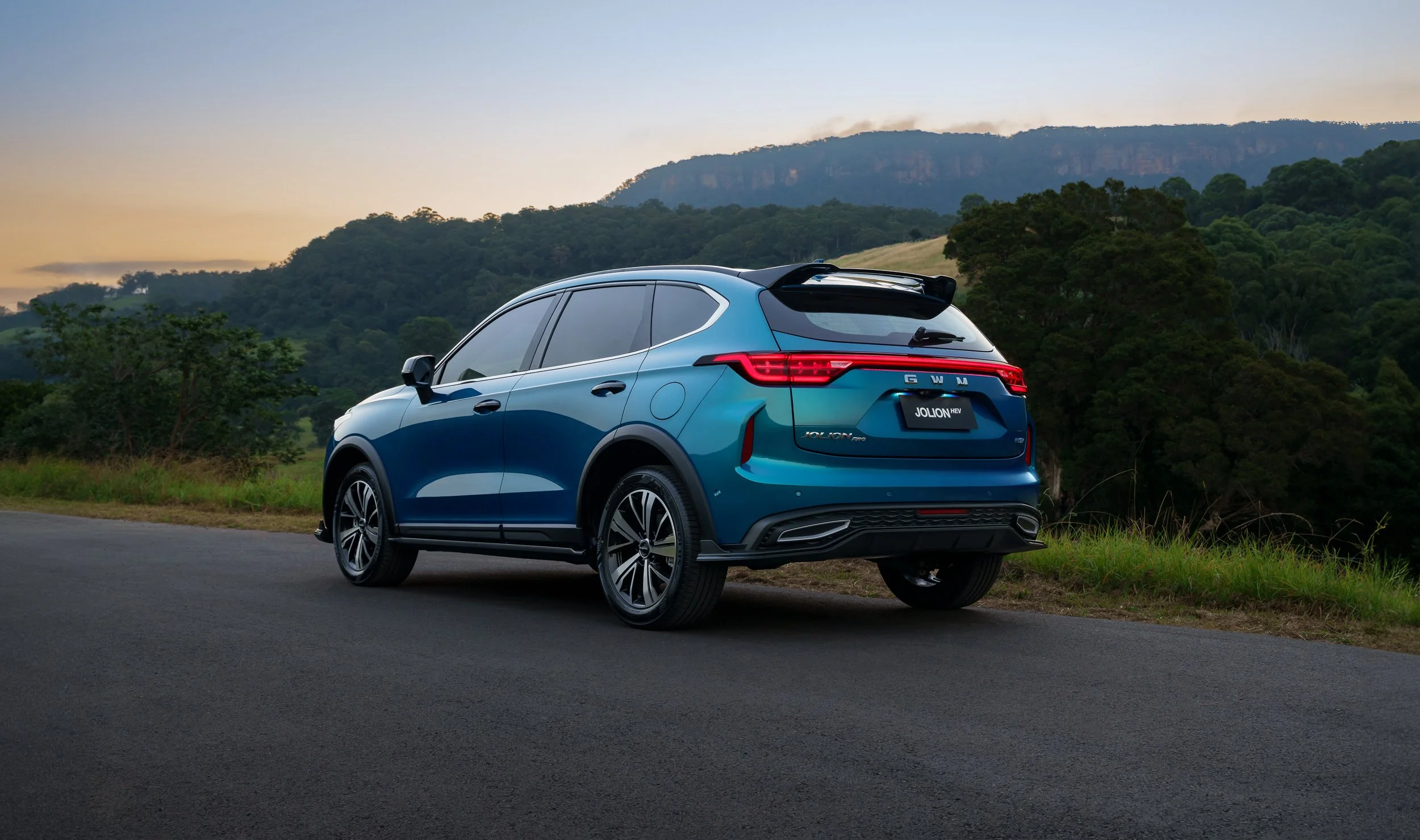 Blue SUV parked on the side of a rural road during sunset with mountains and forest in the background.