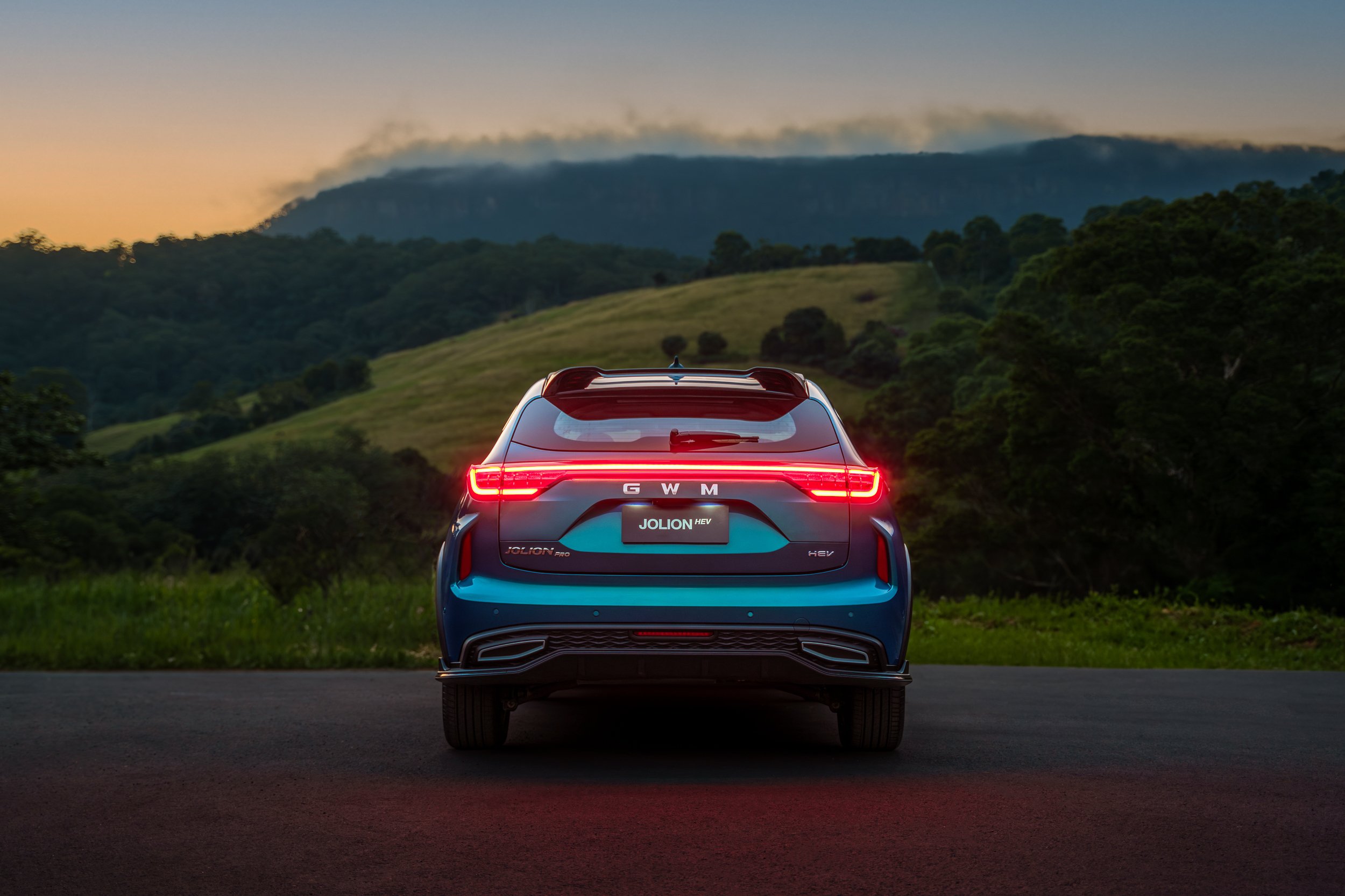 Rear view of a blue electric SUV with illuminated taillights, parked outdoors during dusk, with green hills and trees in the background.