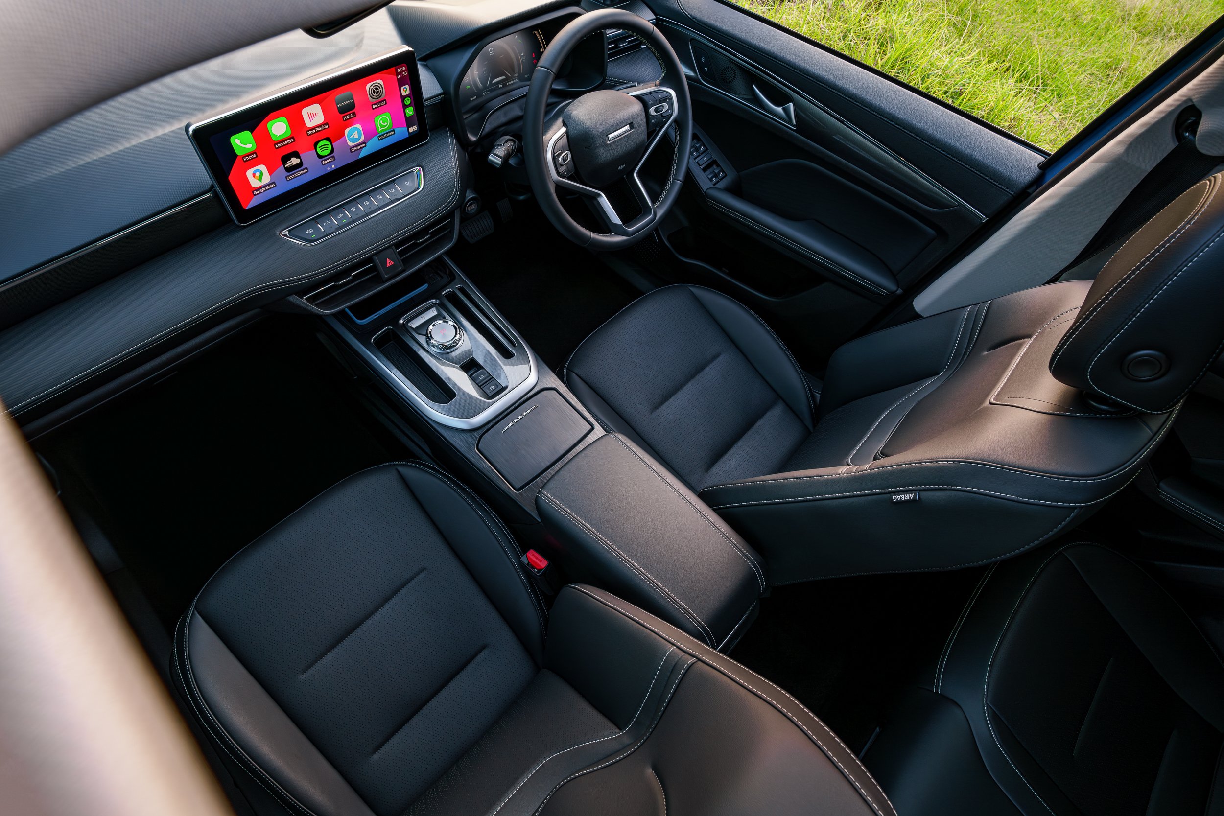 Interior of a modern vehicle showing a steering wheel, gear shift, touchscreen infotainment system displaying various apps, and black leather seating.