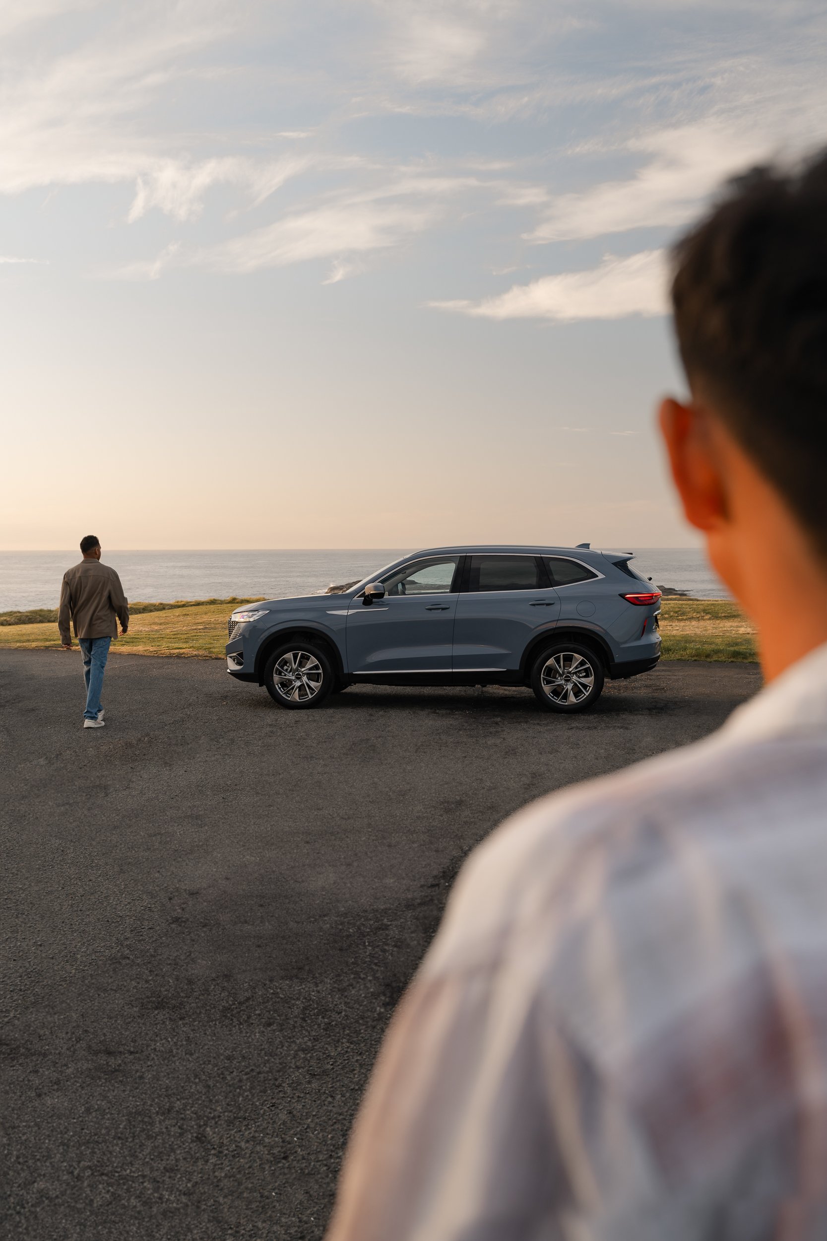 A person with short dark hair is in the foreground on the right side of the image, blurry. In the background, there is a man walking away near a car parked on a roadside with the ocean and sky visible, with some clouds, during sunset or sunrise.