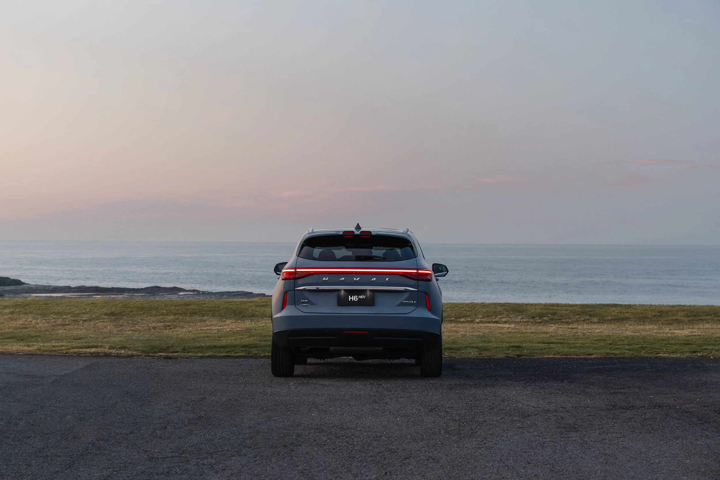 A silver electric vehicle parked on a grassy area near the ocean during dusk with a pastel-colored sky.