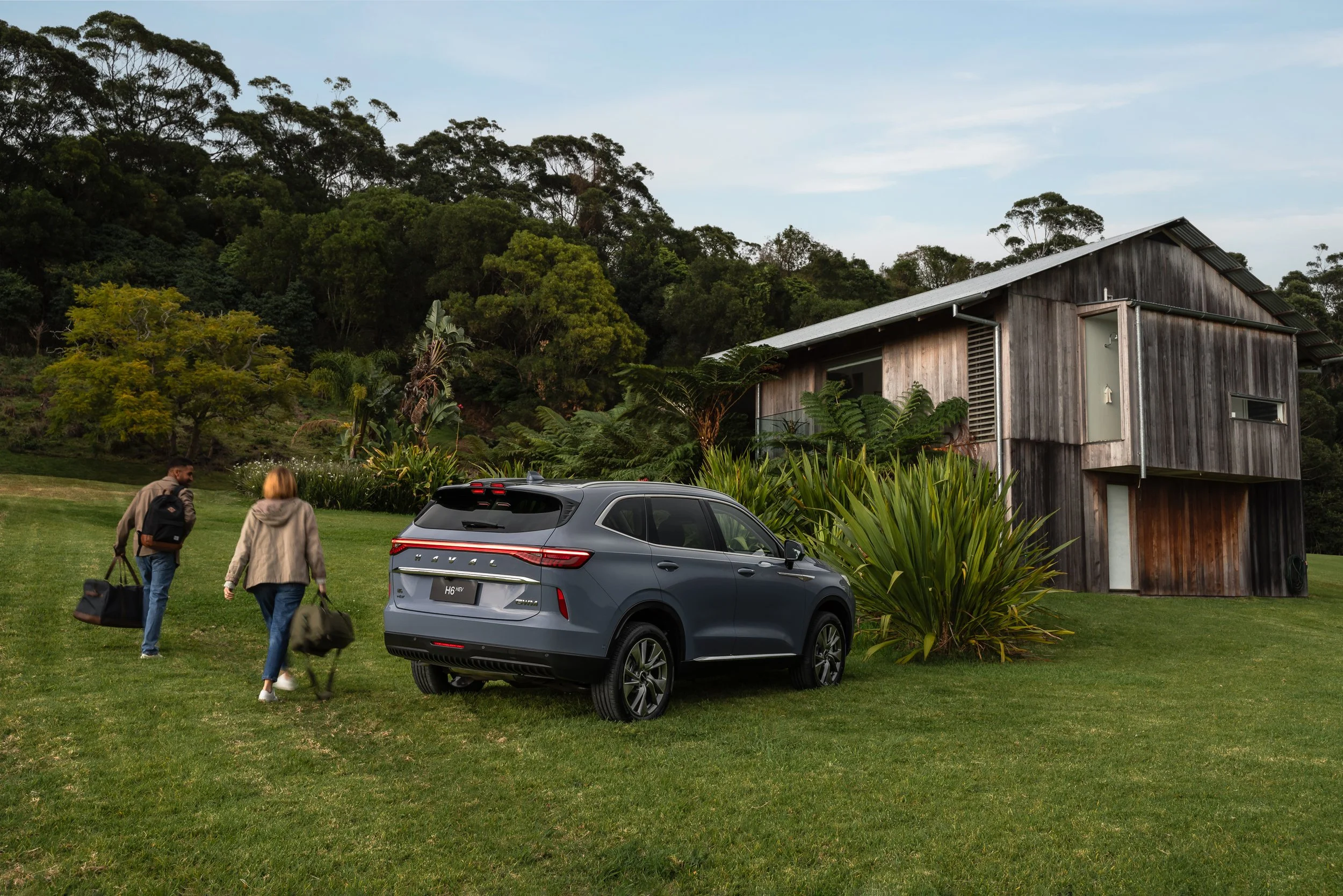 Two people are walking across a grassy area towards a modern wooden house with lush green trees and bushes in the background. A gray SUV is parked nearby.