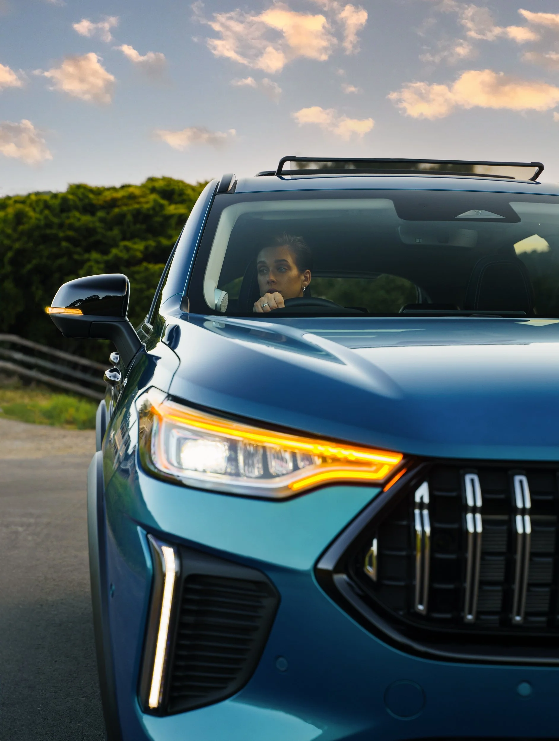 Close-up of the front of a blue luxury SUV, with a woman sitting inside, looking out through the windshield during sunset with a background of green hills and a partly cloudy sky.