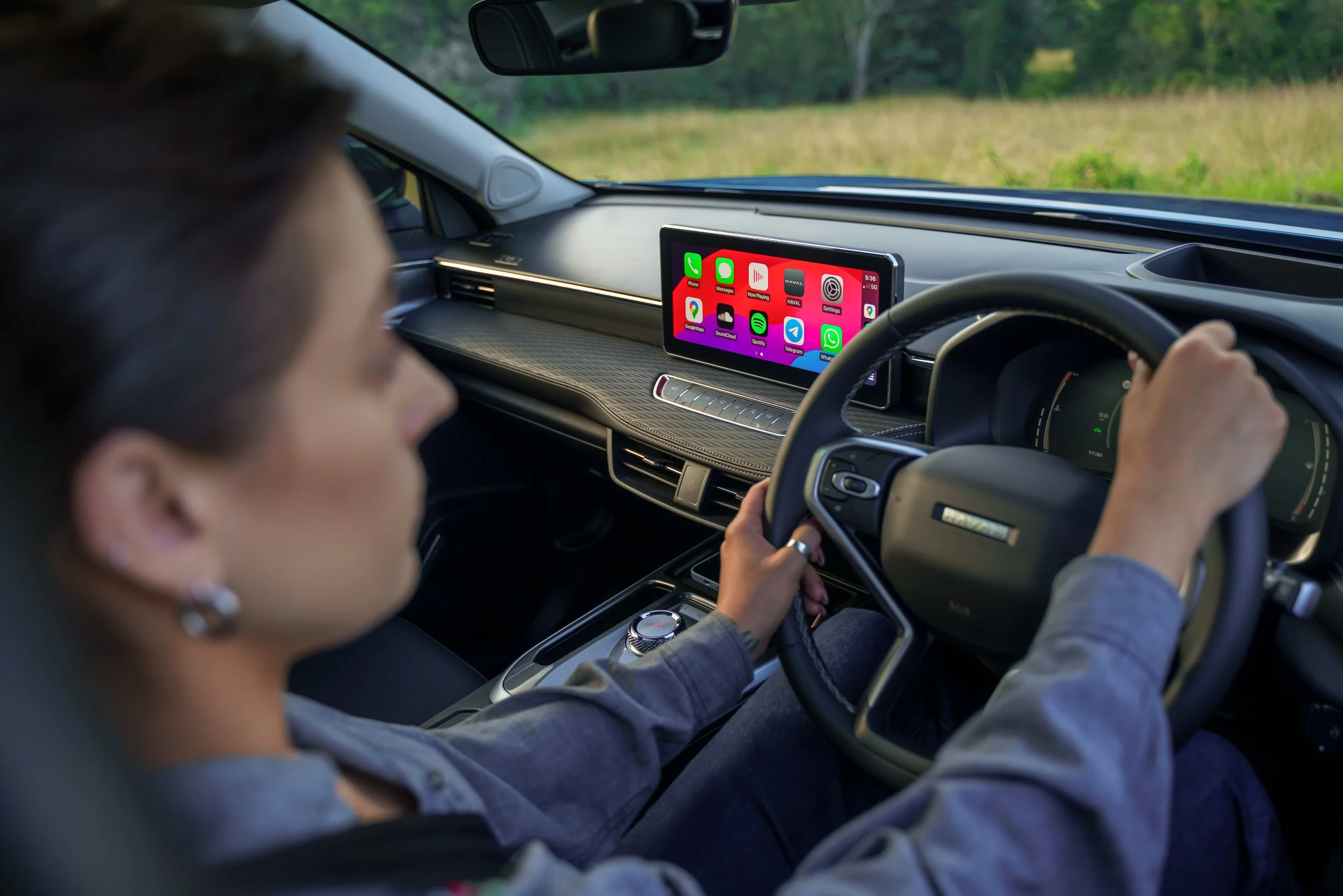 Person driving a car with a large digital display screen on the dashboard showing various app icons, including Phone, Messages, Spotify, and WhatsApp, with a view of greenery outside through the windshield.