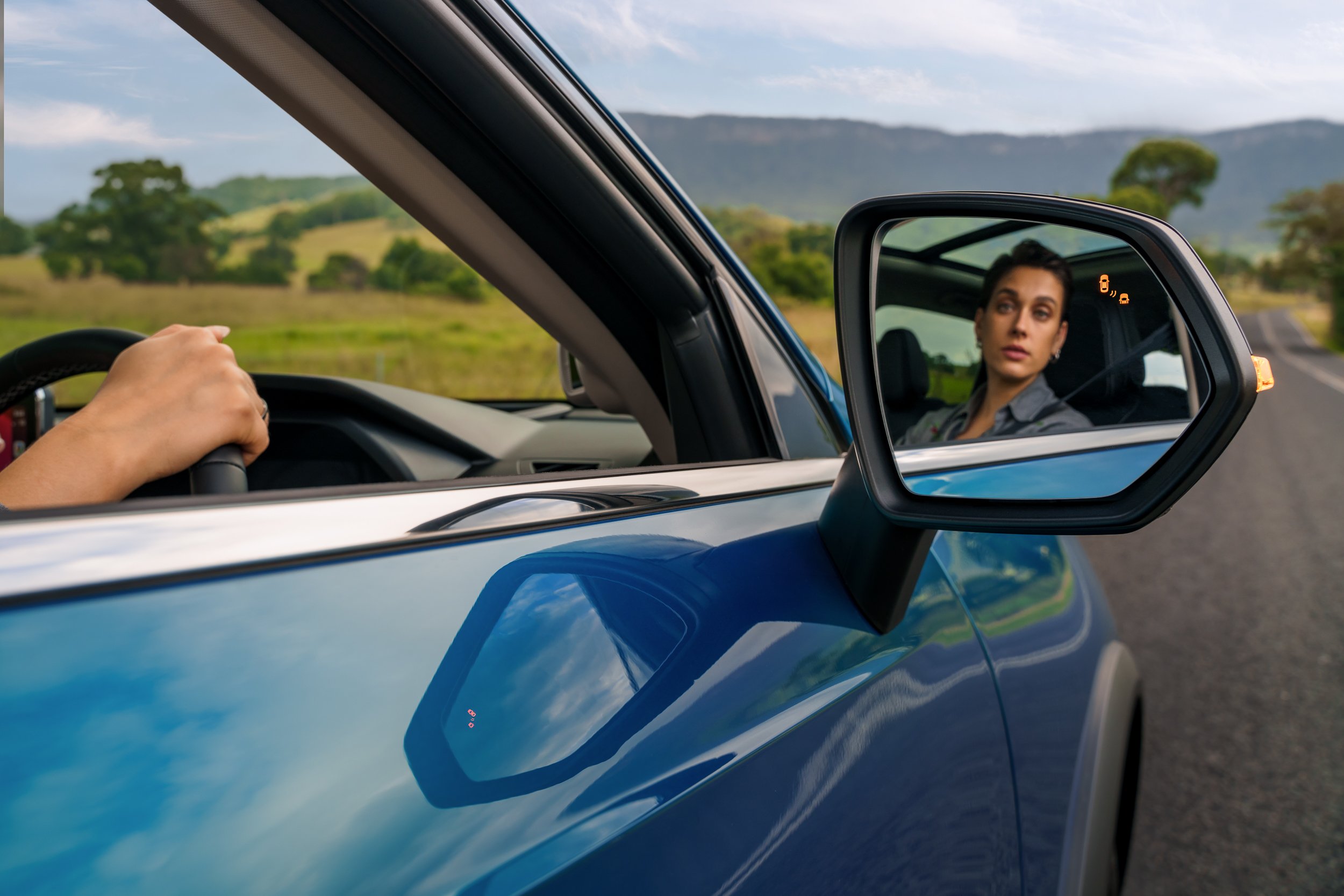A woman looking at her reflection in the side mirror of a blue vehicle, while a person is driving the vehicle in a rural area with green trees and hills in the background.