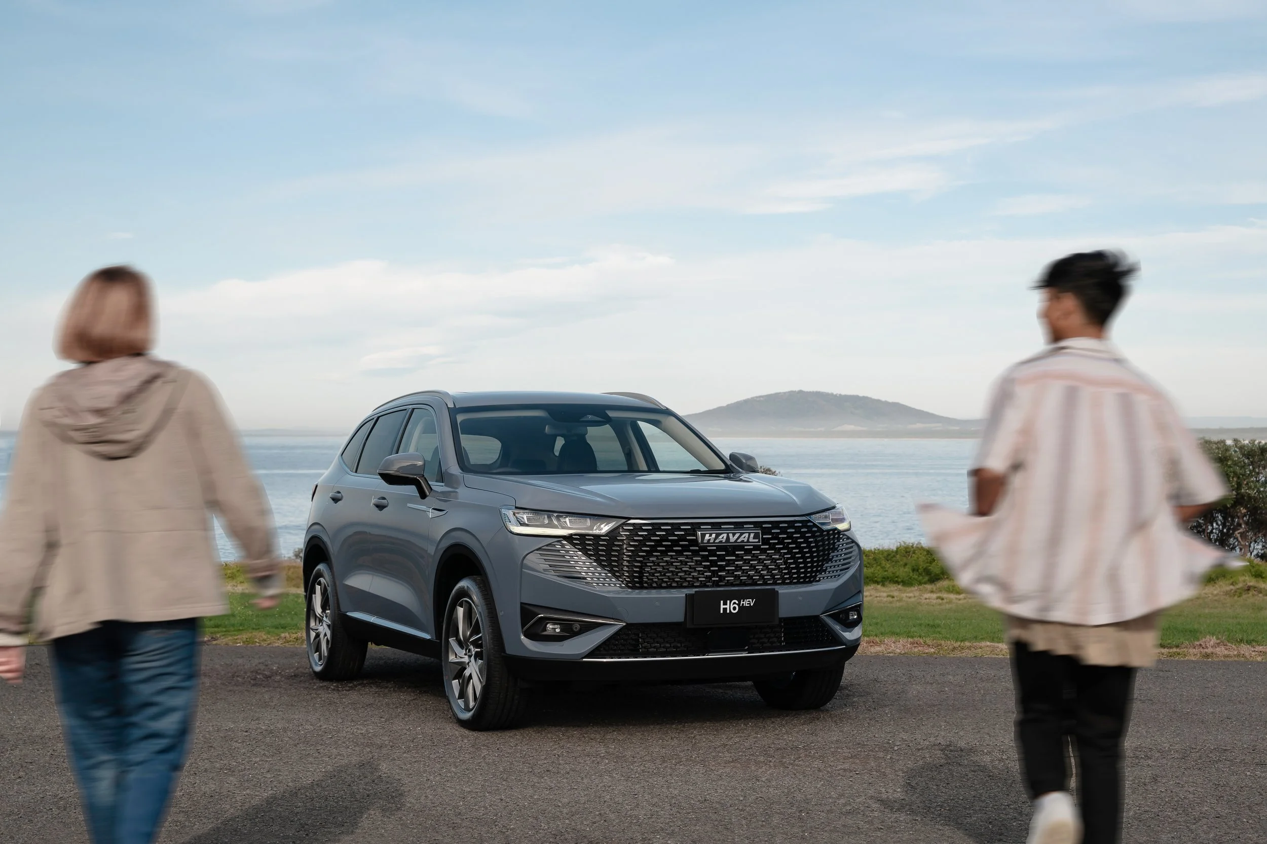 A grey Haval H6 HEV SUV parked outdoors near a body of water with mountains in the background. Two people, a woman and a man, walk past the vehicle. The scene appears to be at a scenic lookout or park by the water.