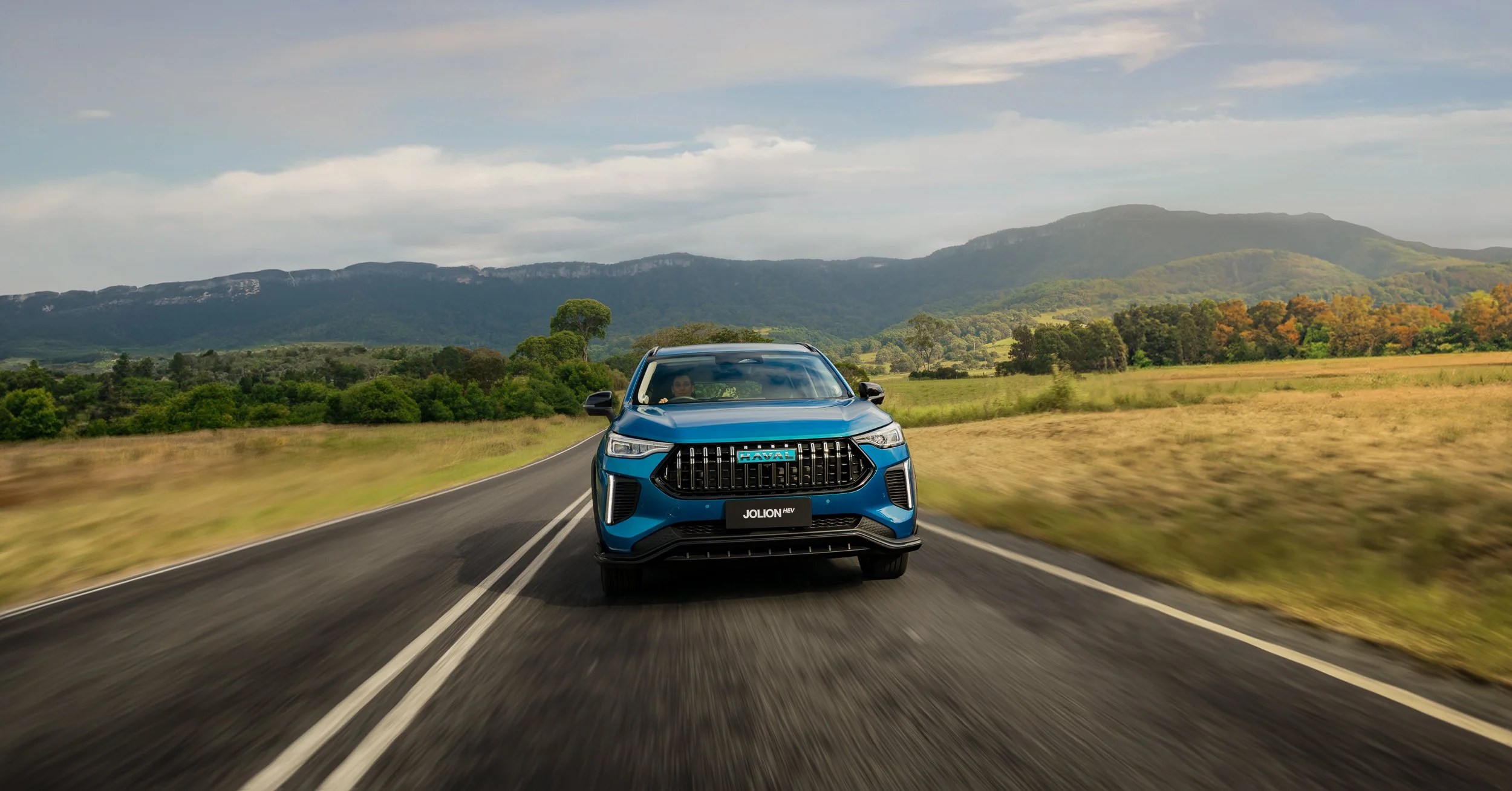 A blue Haval Jolion HEV SUV driving on a rural two-lane road with mountains and green fields in the background.