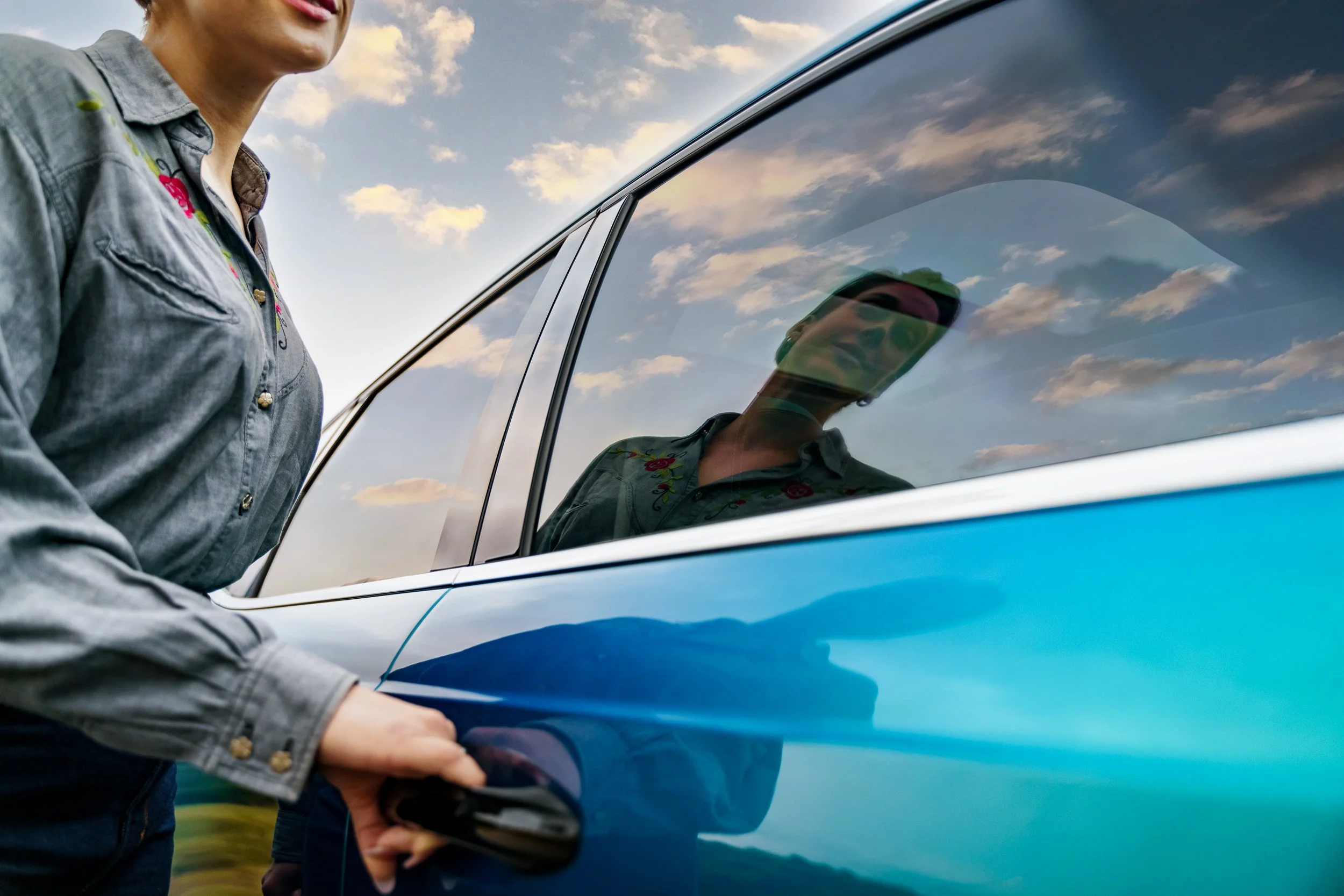 A person with short hair, wearing a gray denim jacket with a floral embroidery shirt, is opening a classic blue car's door. A reflection of their face and sunglasses can be seen in the car window. The sky is partly cloudy with soft light.
