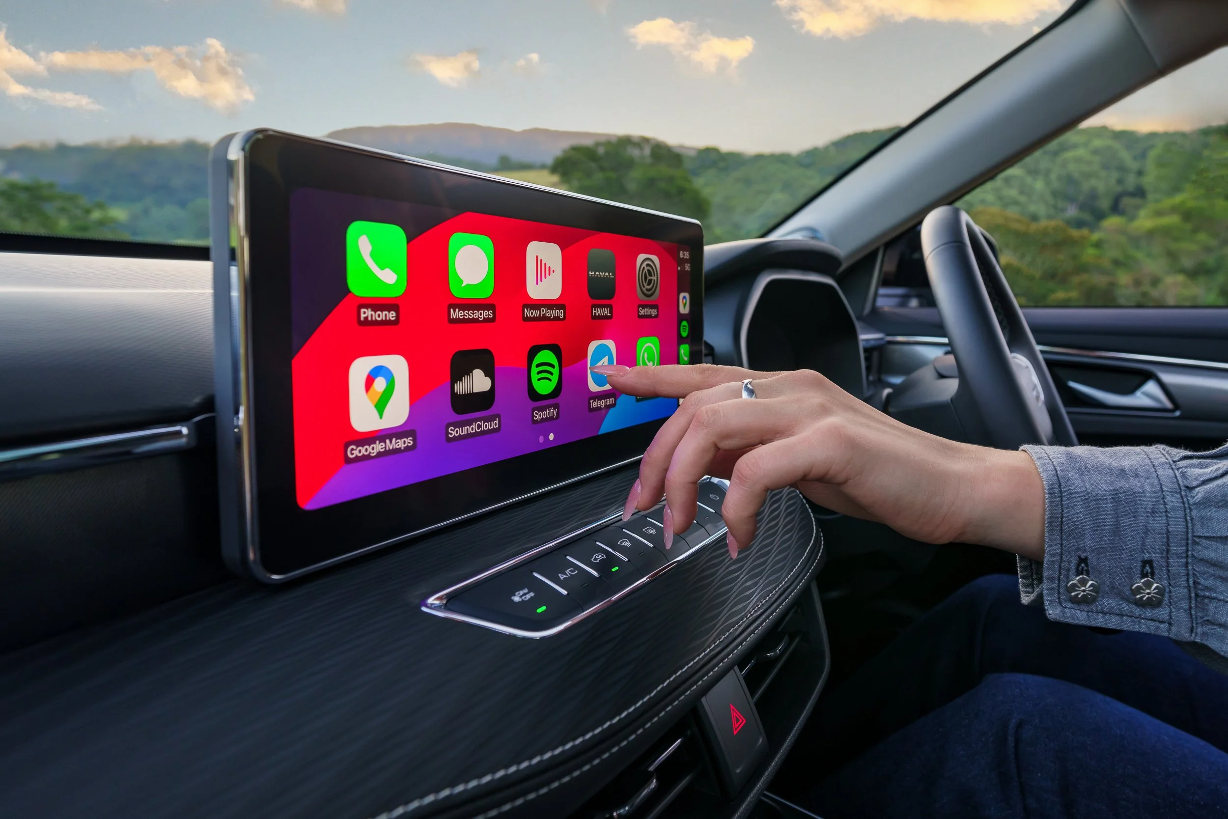 Dashboard navigation screen inside a car with a person's hand interacting, showing app icons for phone, messages, music, navigation, and communication apps against a scenic landscape background.