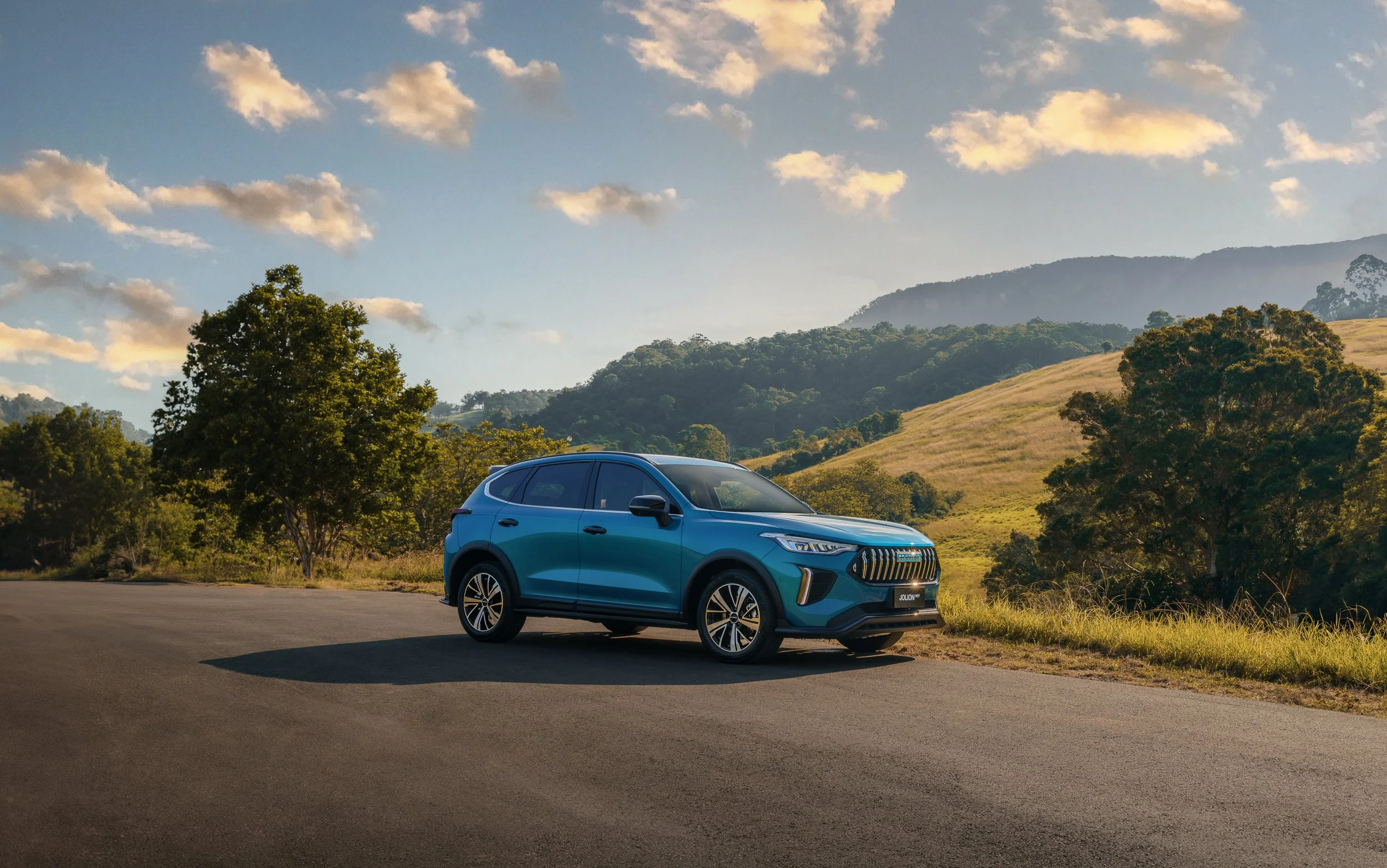 Blue SUV parked on a paved area near grassy ground surrounded by trees, hills, and a partly cloudy sky during daytime.