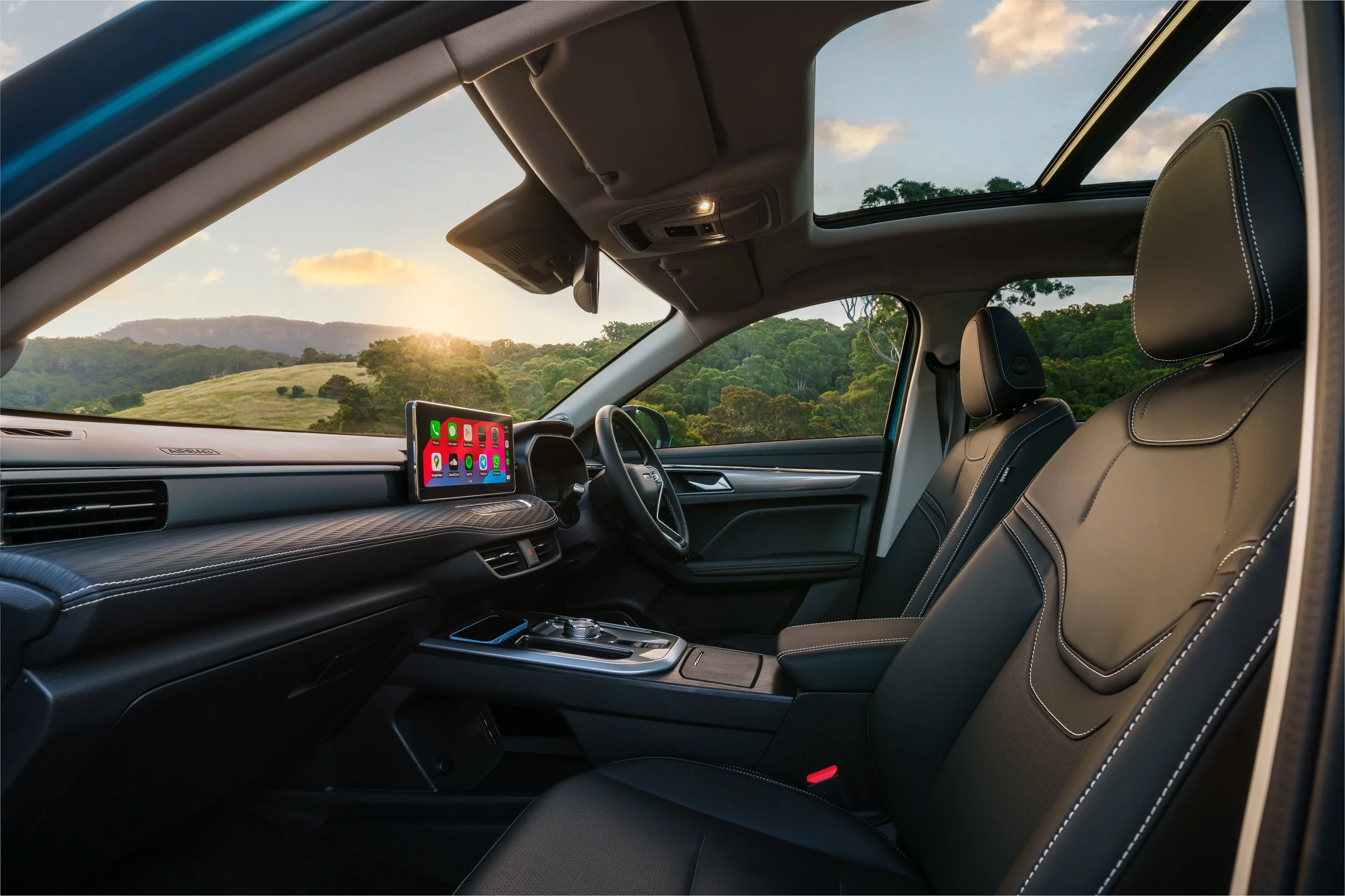 View of the interior of a modern vehicle with black seats, a dashboard with a mounted smartphone displaying app icons, and a scenic landscape of green hills and trees outside the windshield, illuminated by the setting sun.