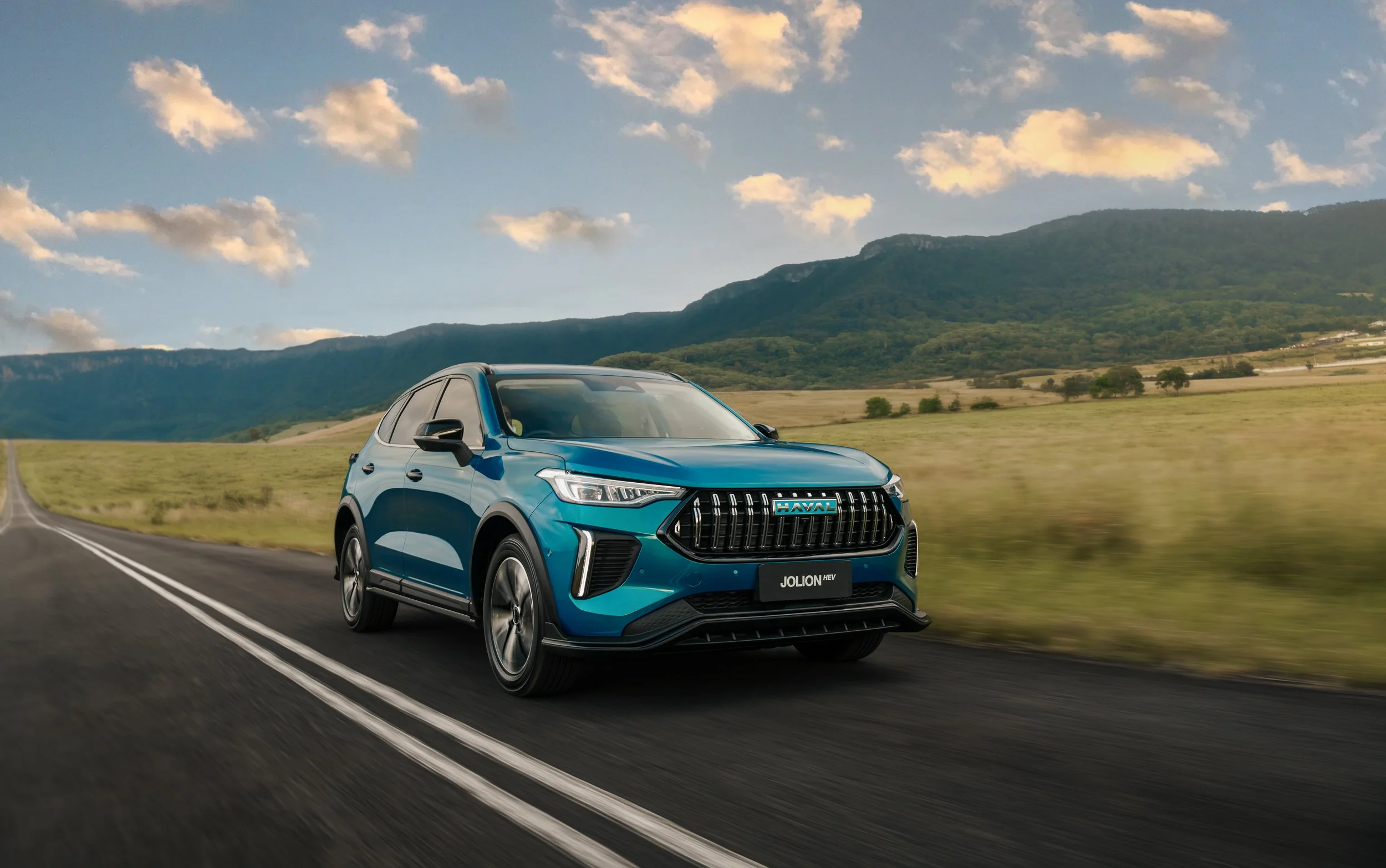 A blue Haval Jolion HEV SUV driving on a scenic open road with mountains and a partly cloudy sky in the background.