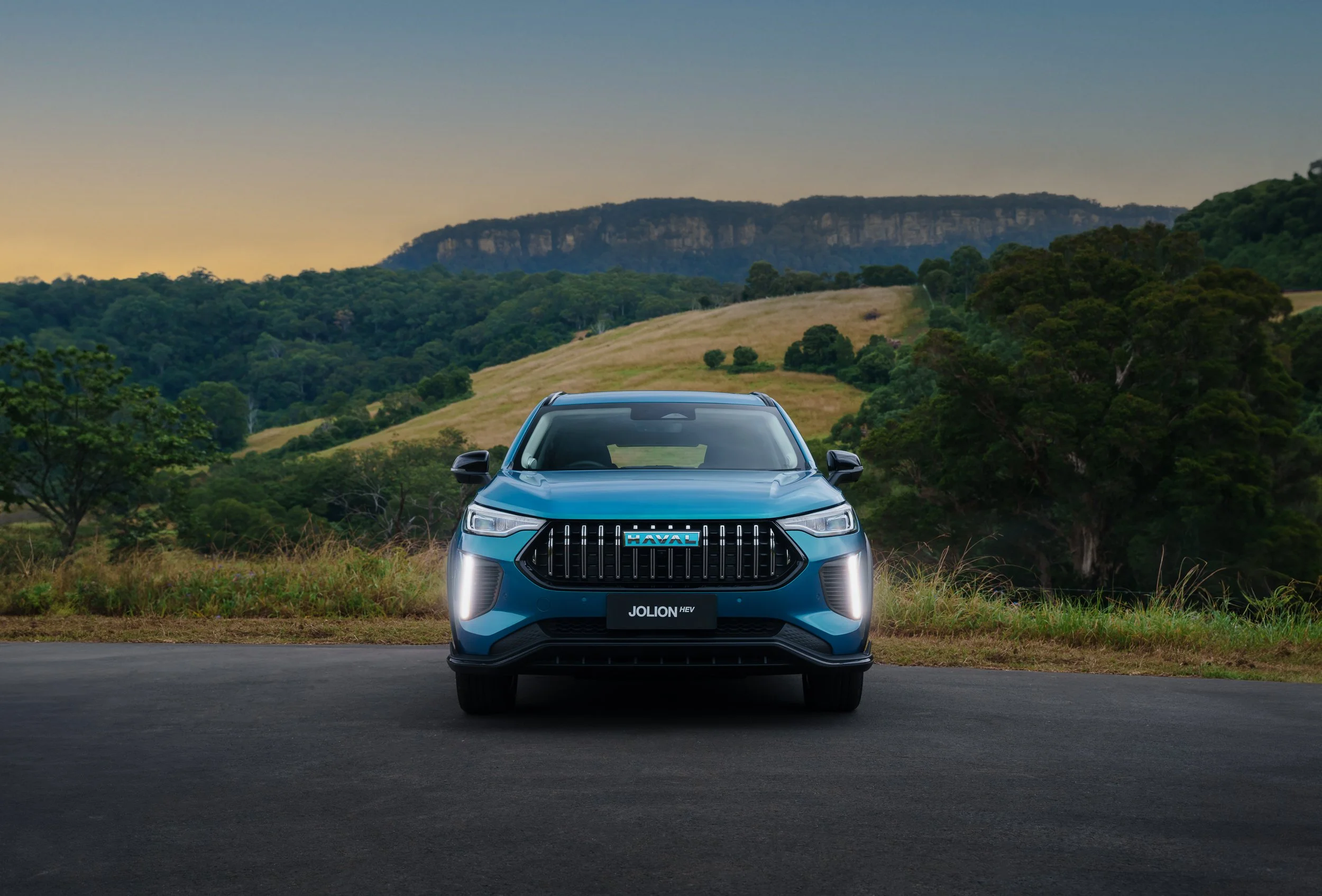 Blue electric SUV parked on a road with a scenic backdrop of rolling hills, green trees, and a mountain at sunset.
