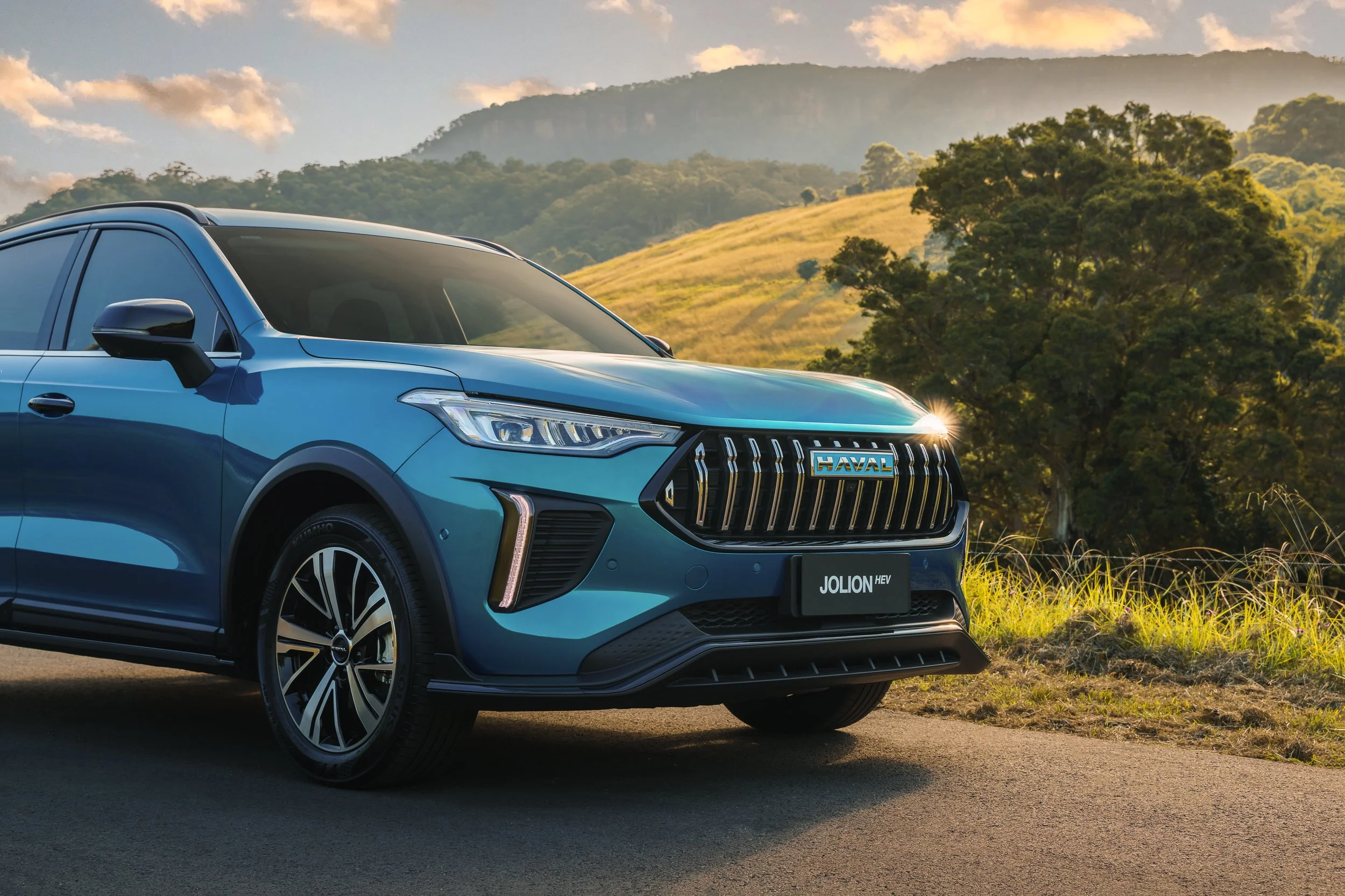 A blue Haval Jolion HEV SUV parked on the side of a paved road, with mountains, green trees, and partly cloudy sky in the background during sunset.