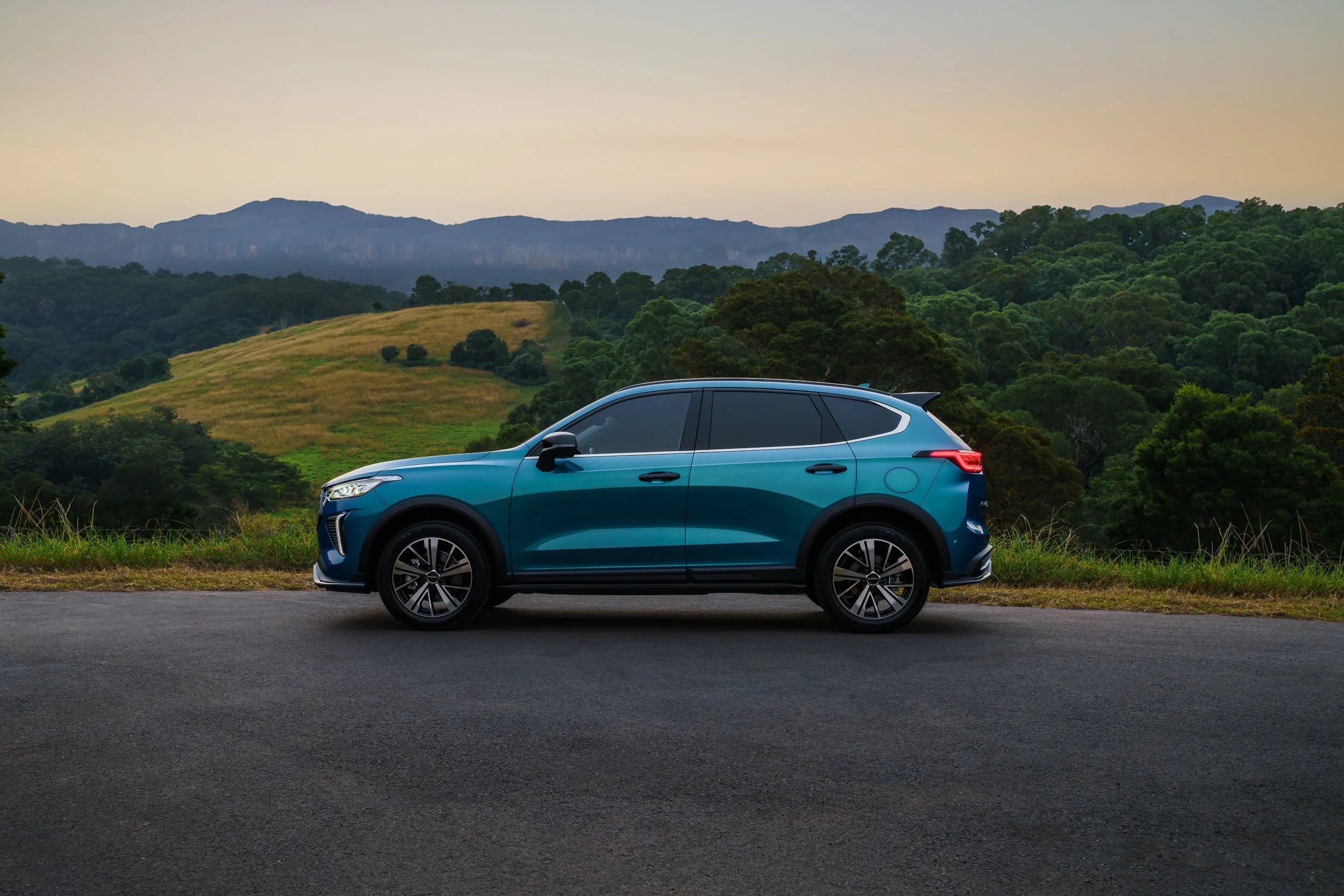Blue SUV parked on a paved road with green forested hills and mountains in the background during daytime.