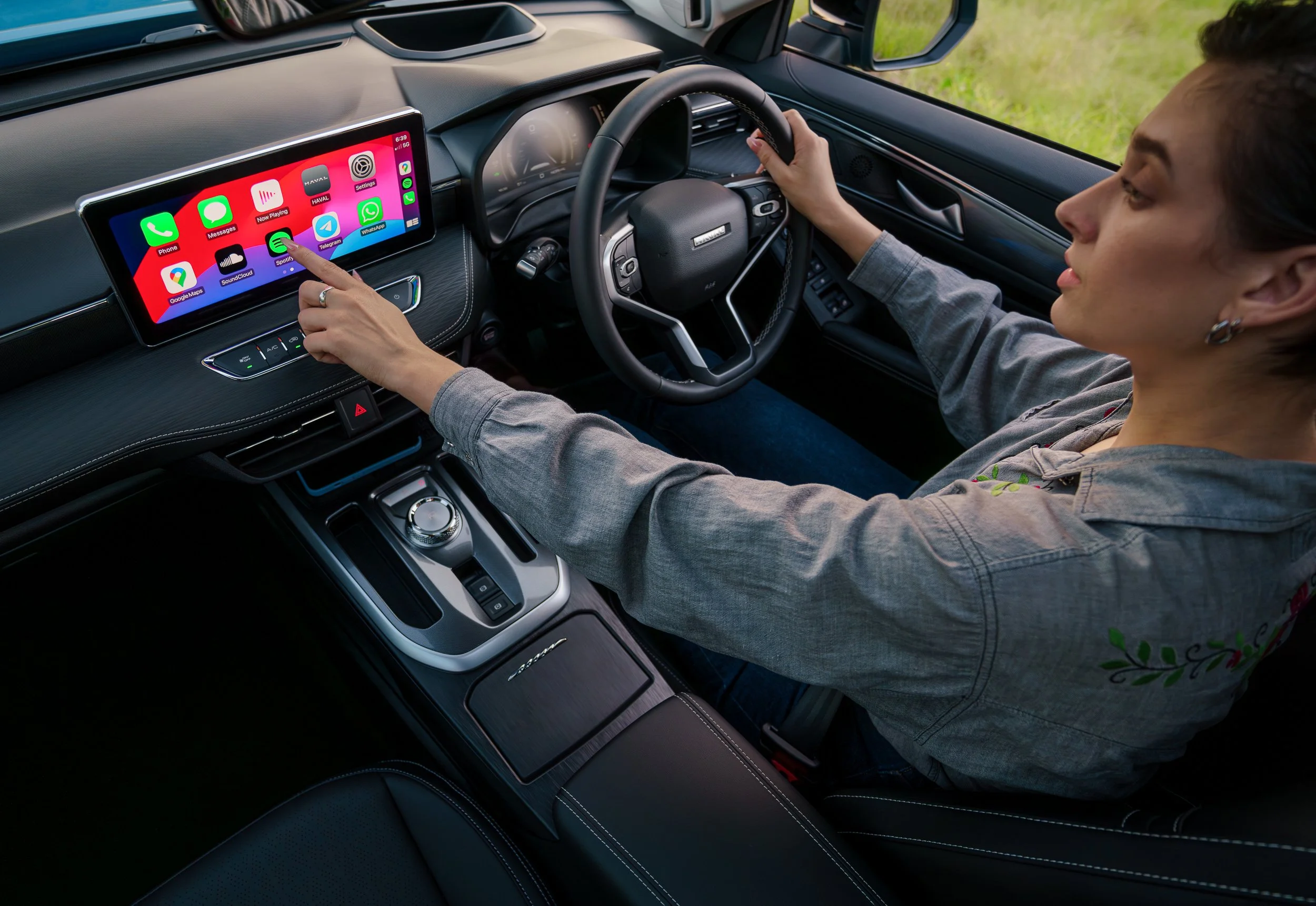 A woman sitting in the driver's seat of a car, using a touchscreen infotainment system. The screen displays various app icons, including Phone, Messages, Spotify, WhatsApp, and Google Maps. The interior of the car features a modern dashboard and stee