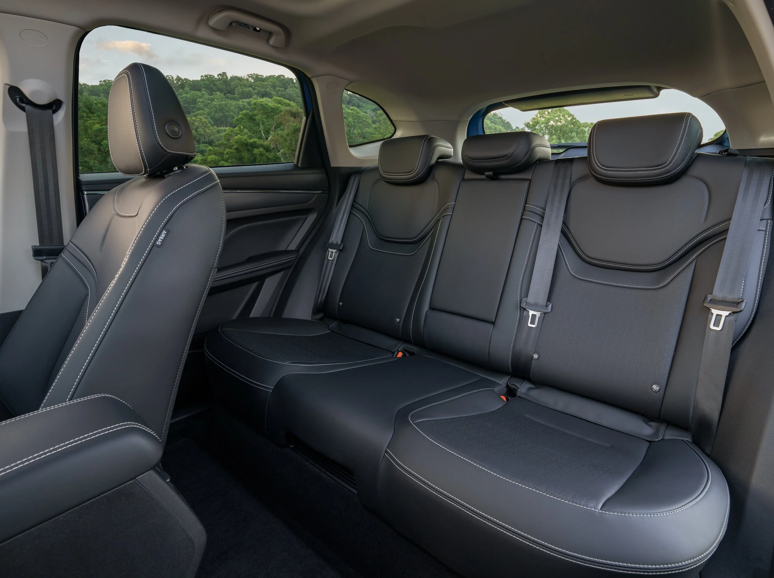 The interior of a vehicle showing black leather seats with white stitching, headrests, and a view of greenery outside through the windows.