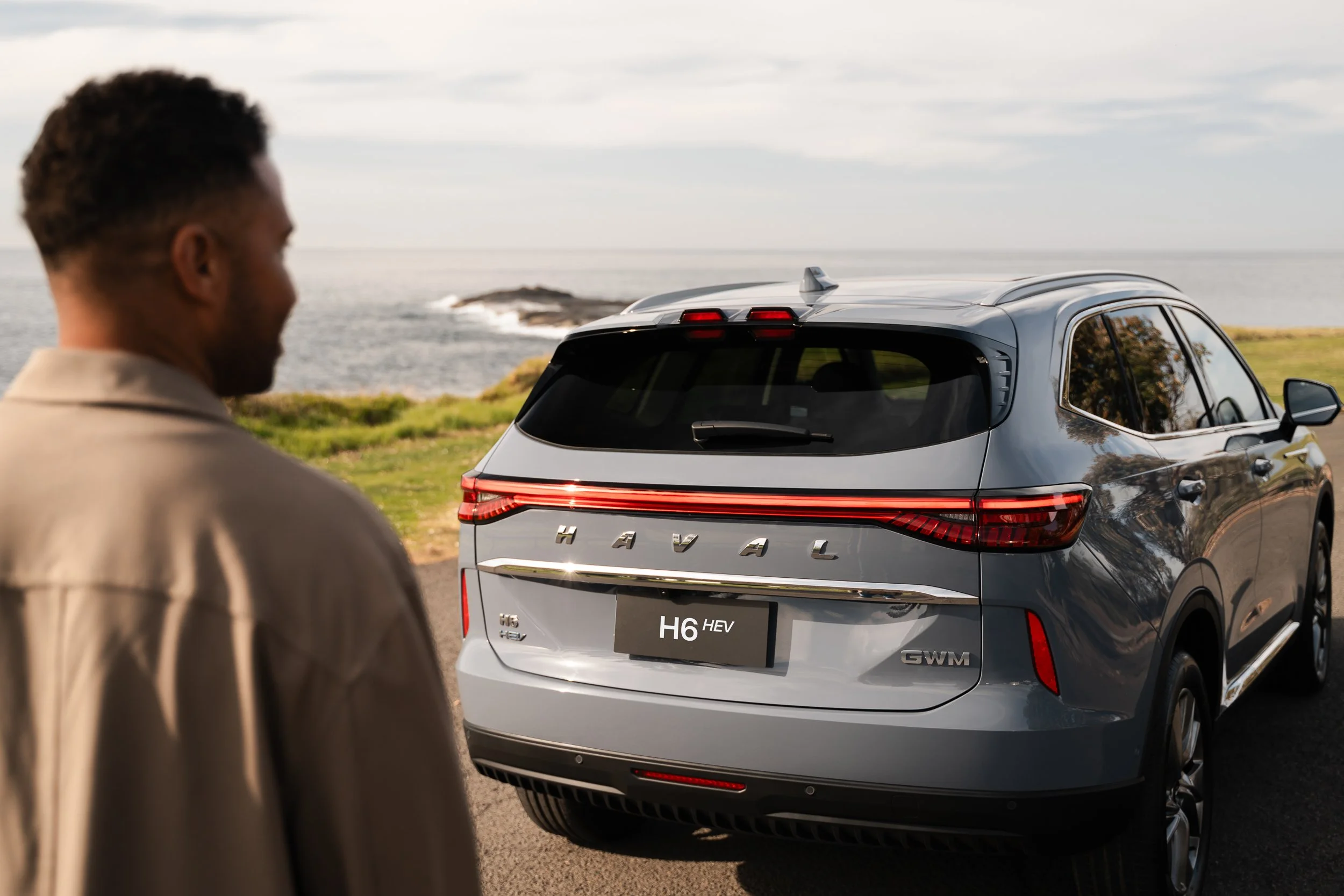 A man with short dark hair looking at a gray Chevrolet H6 HEV SUV by the coast, with ocean and cloudy sky in the background.