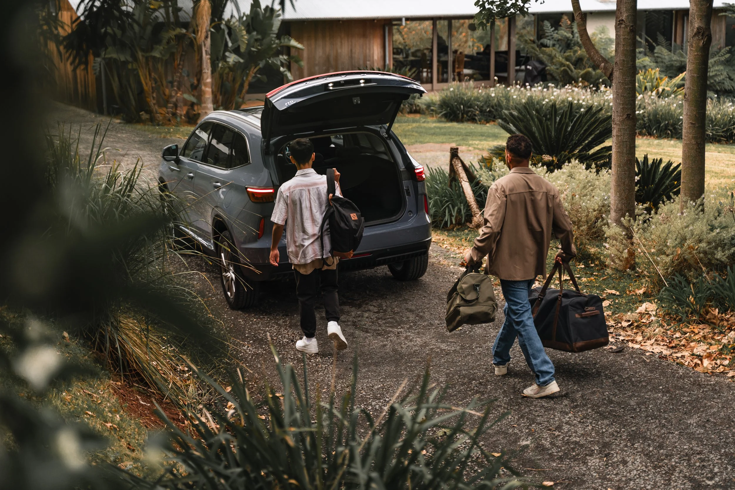 Two men are walking towards a gray SUV with an open trunk filled with bags and luggage in a garden with trees and plants. One man has short dark hair, is wearing a jacket and jeans, and is carrying a duffel bag. The other man is wearing a loose shirt