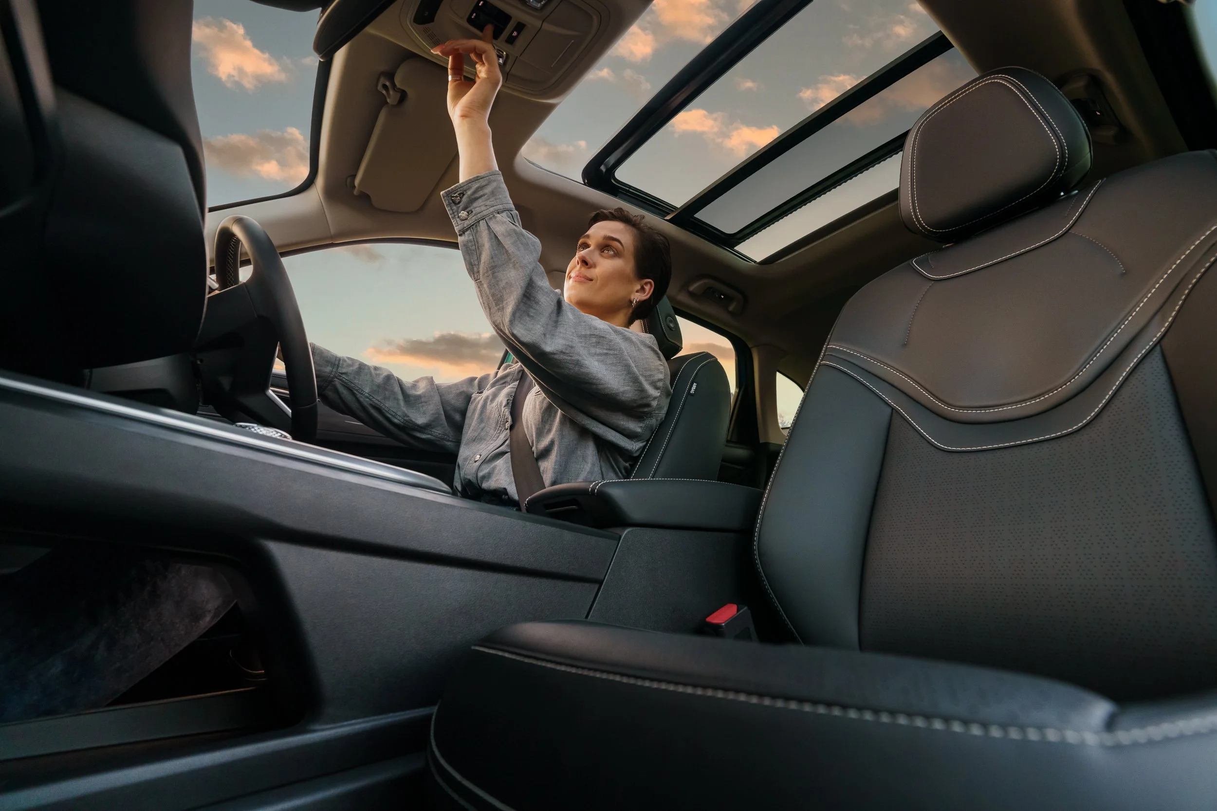 A woman sitting in the driver’s seat of a car with a sunroof, reaching up to adjust the ceiling controls, during sunset or sunrise with clouds visible through the sunroof.