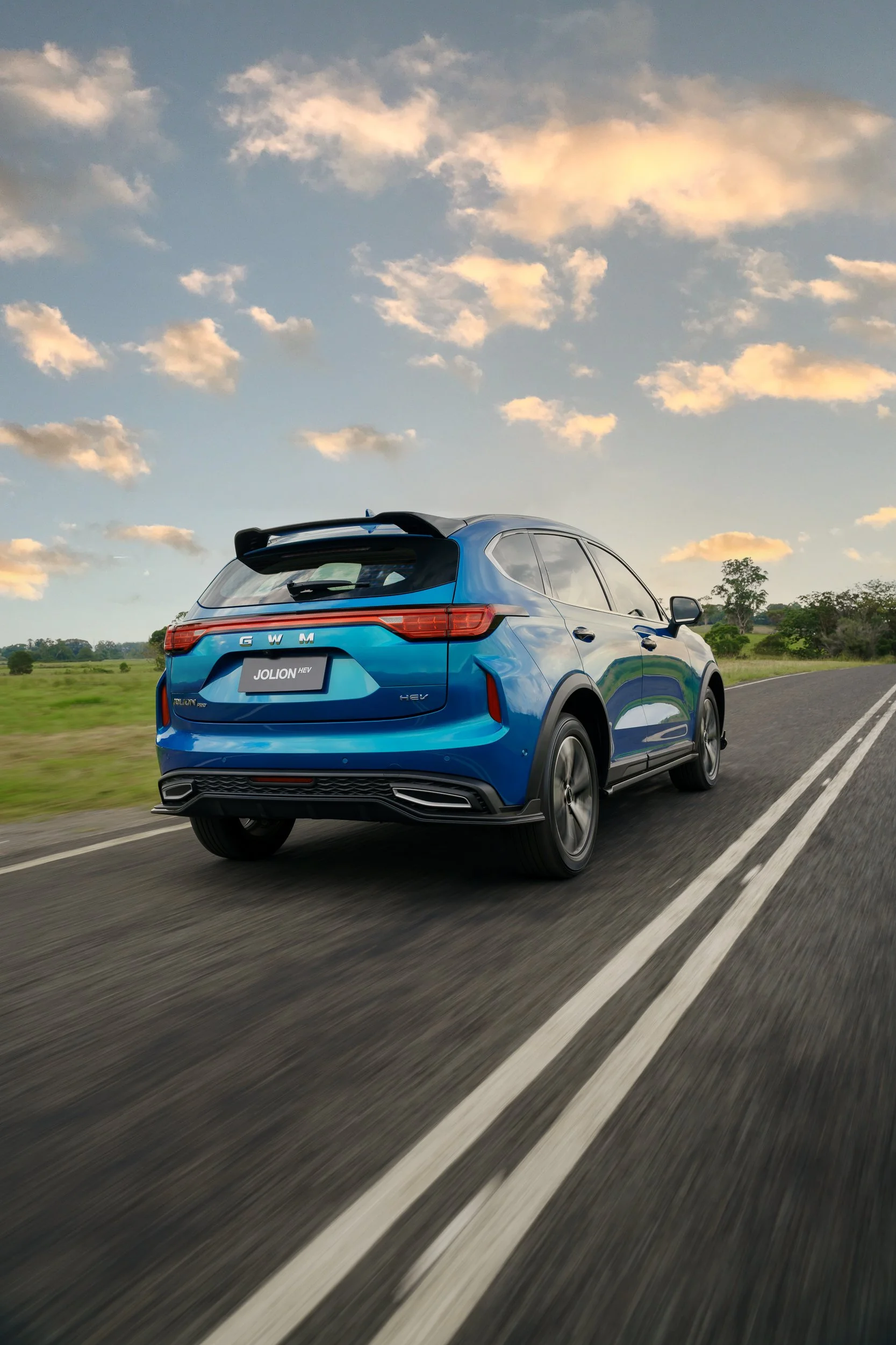 A blue GWM Jolion HEV SUV driving on a two-lane road with green fields and trees in the background, under a sky with scattered clouds during sunset.