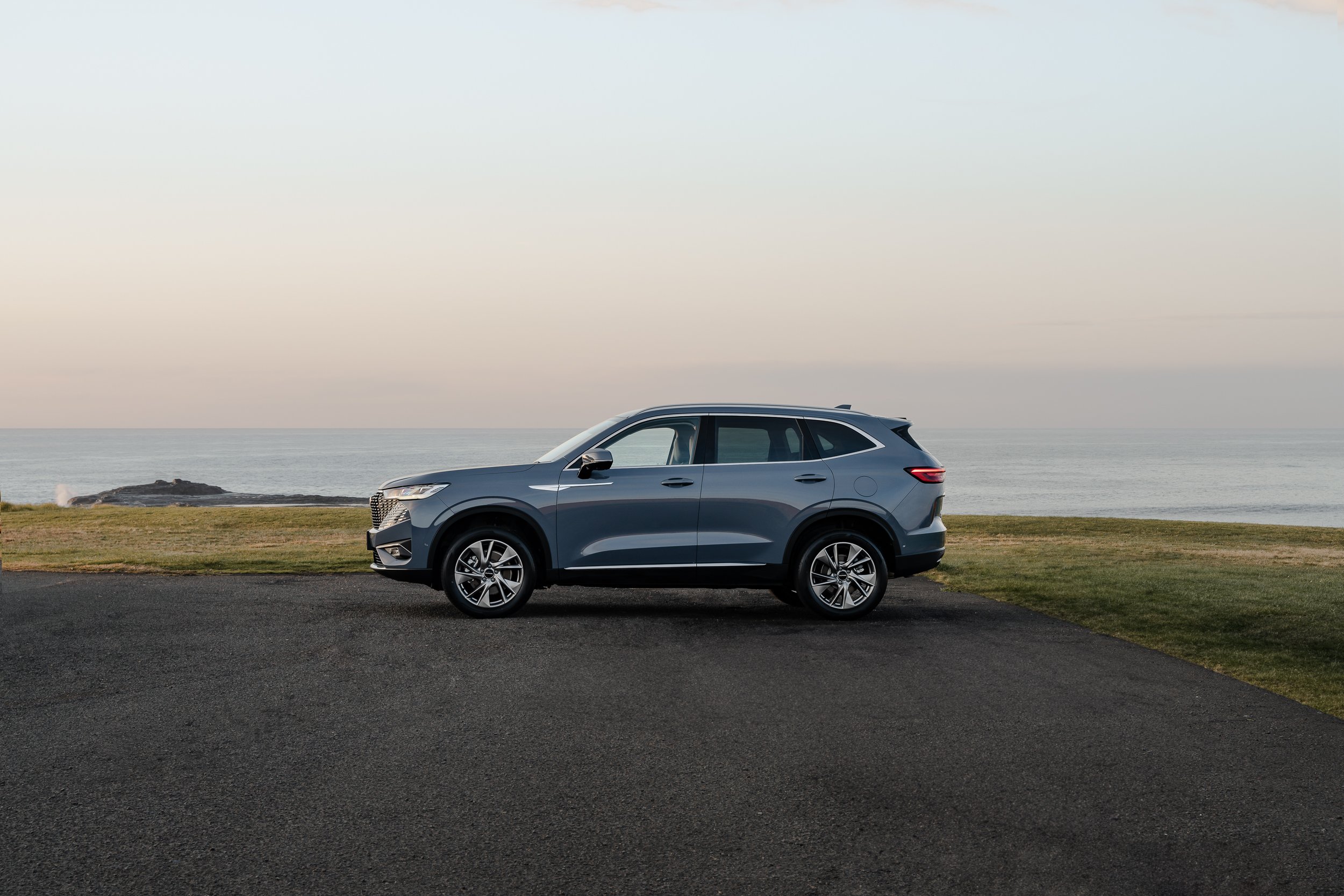 A gray SUV parked on a paved road near the coast with grass and ocean in the background during daytime.