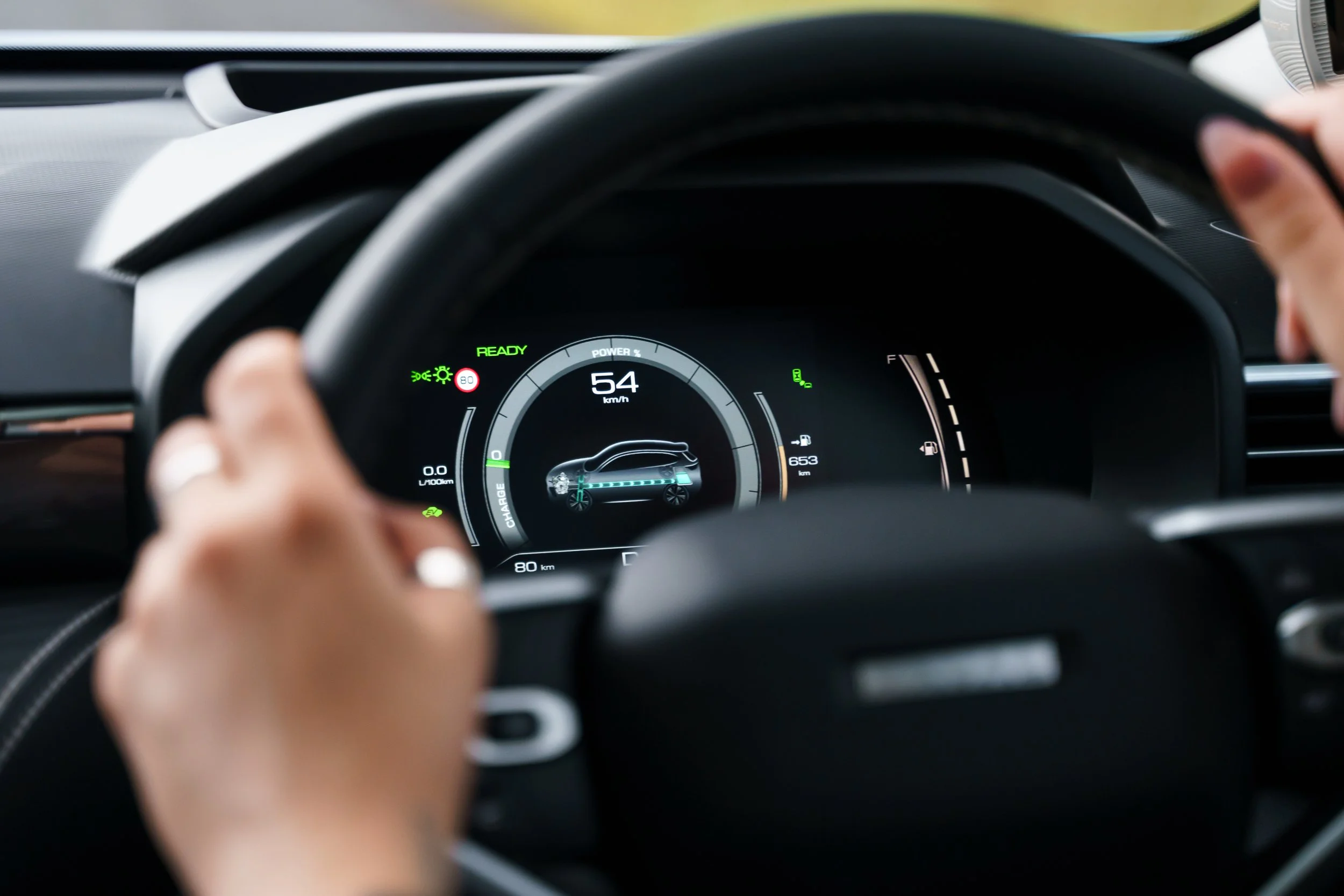 Close-up of a digital car dashboard displaying 54 km/h speed, with a person's hand on the steering wheel. The dashboard shows various indicators, including a green 'READY' signal and an electric vehicle icon.