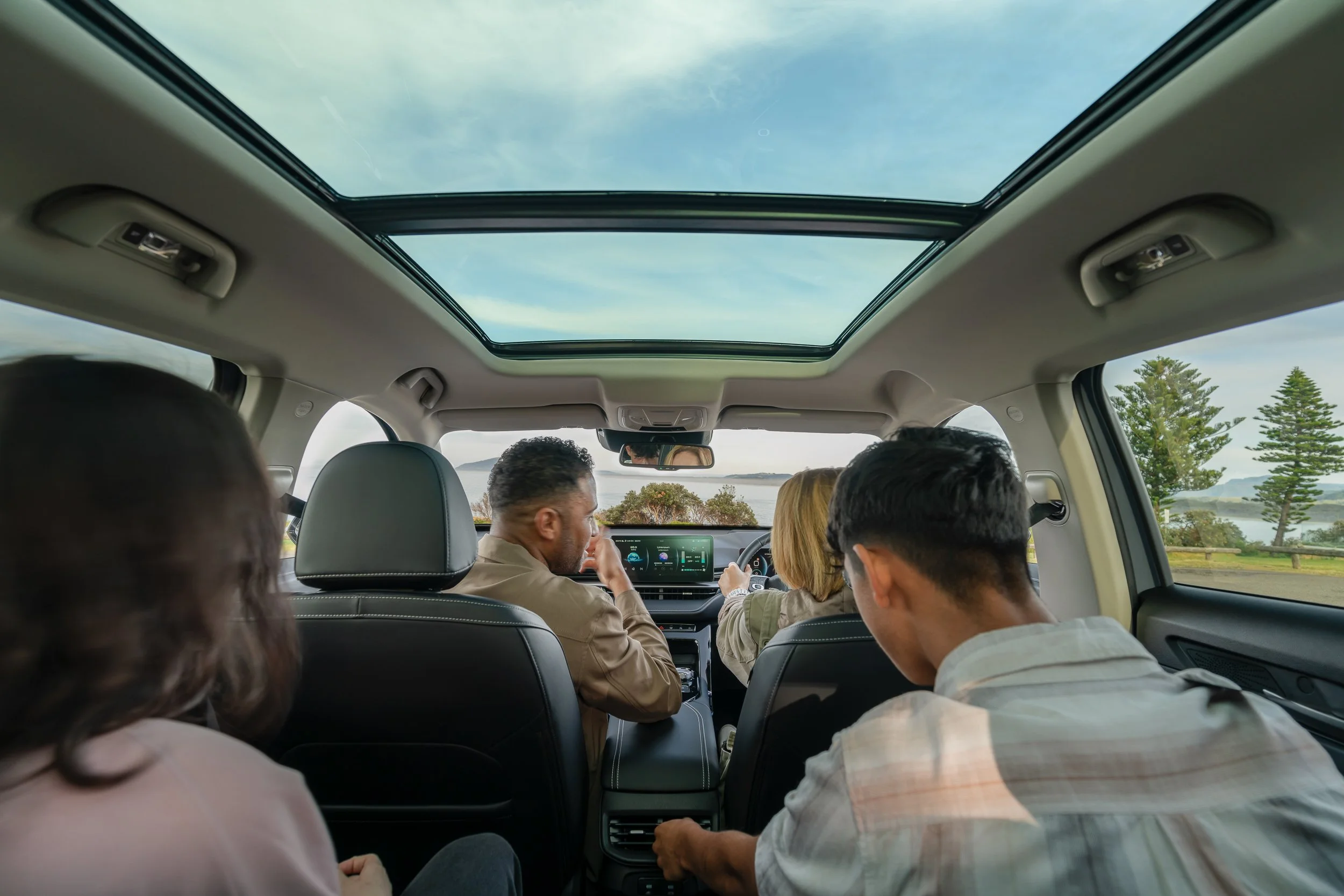Interior of a car with several passengers, including a man with gray hair and a woman with blonde hair, with a panoramic sunroof showing a cloudy sky and trees outside.