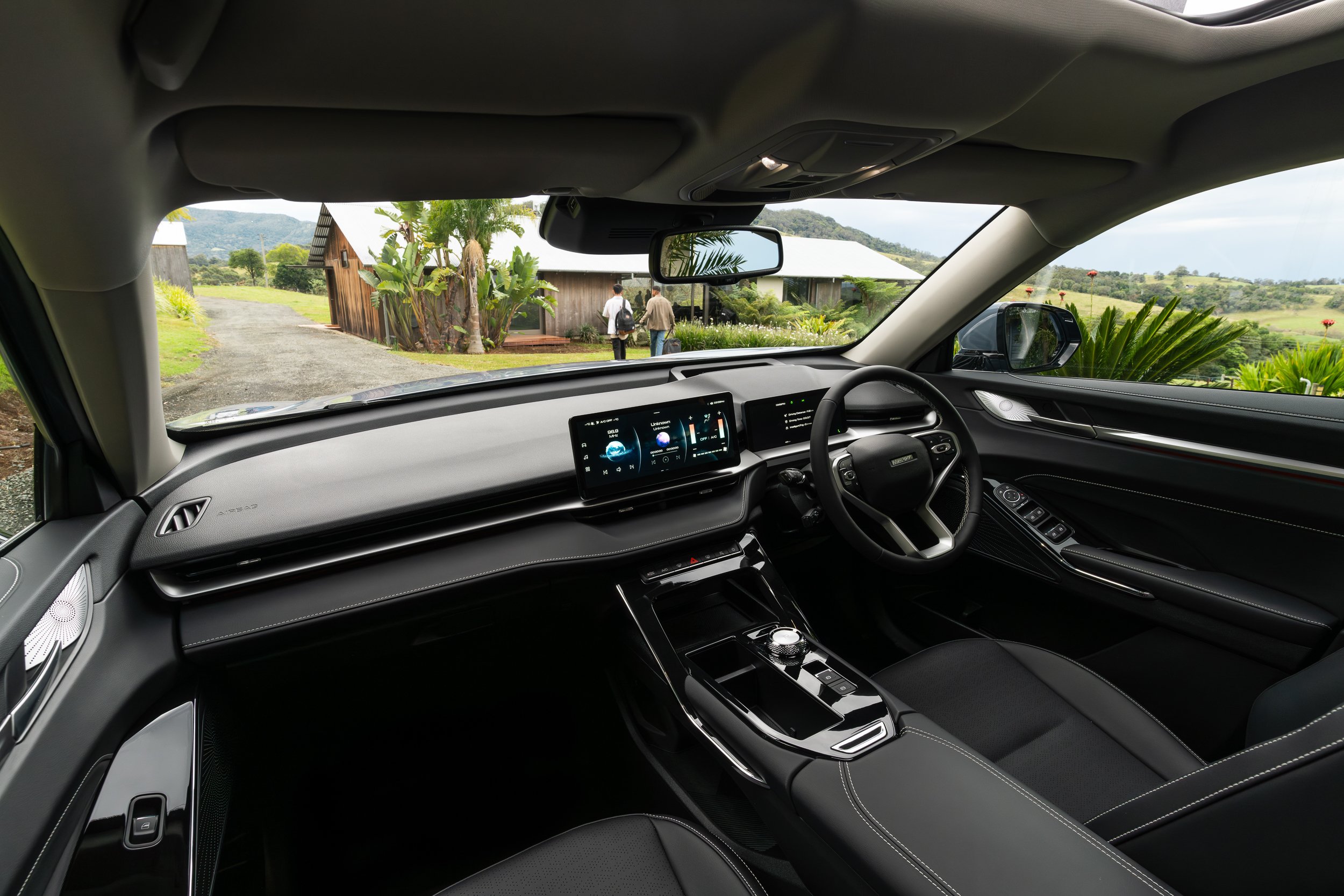 The interior of a modern electric vehicle showing a sleek dashboard with digital displays, steering wheel, and center console, with a view of a rural landscape through the windshield including a dirt road, greenery, and two people walking near a wood