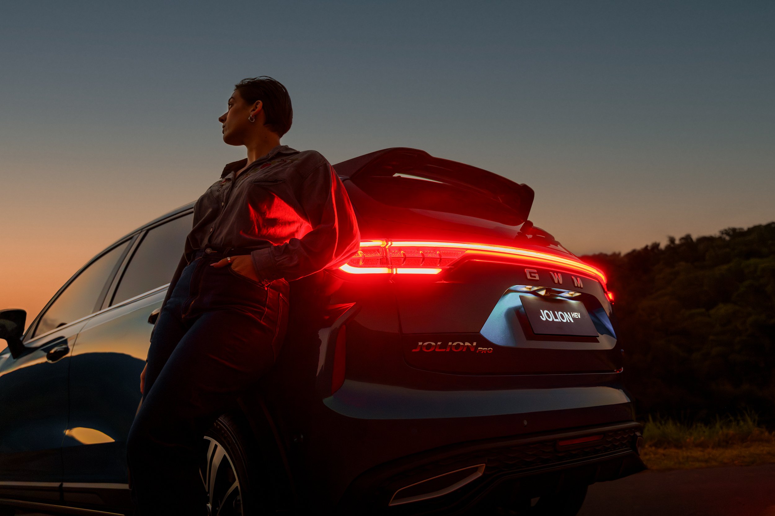 A woman with short dark hair, wearing earrings and a denim jacket, stands beside a black SUV with illuminated red taillights at sunset, with a backdrop of blue sky and distant hills.