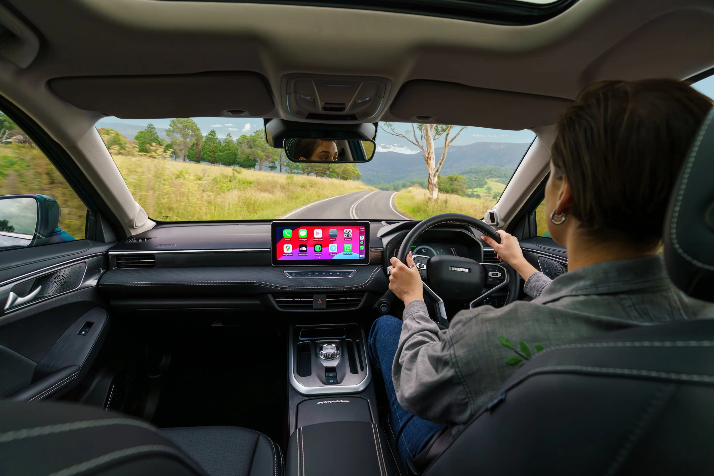 A person driving a car on a scenic rural road with trees and mountains in the distance, using a smartphone mounted on the dashboard.