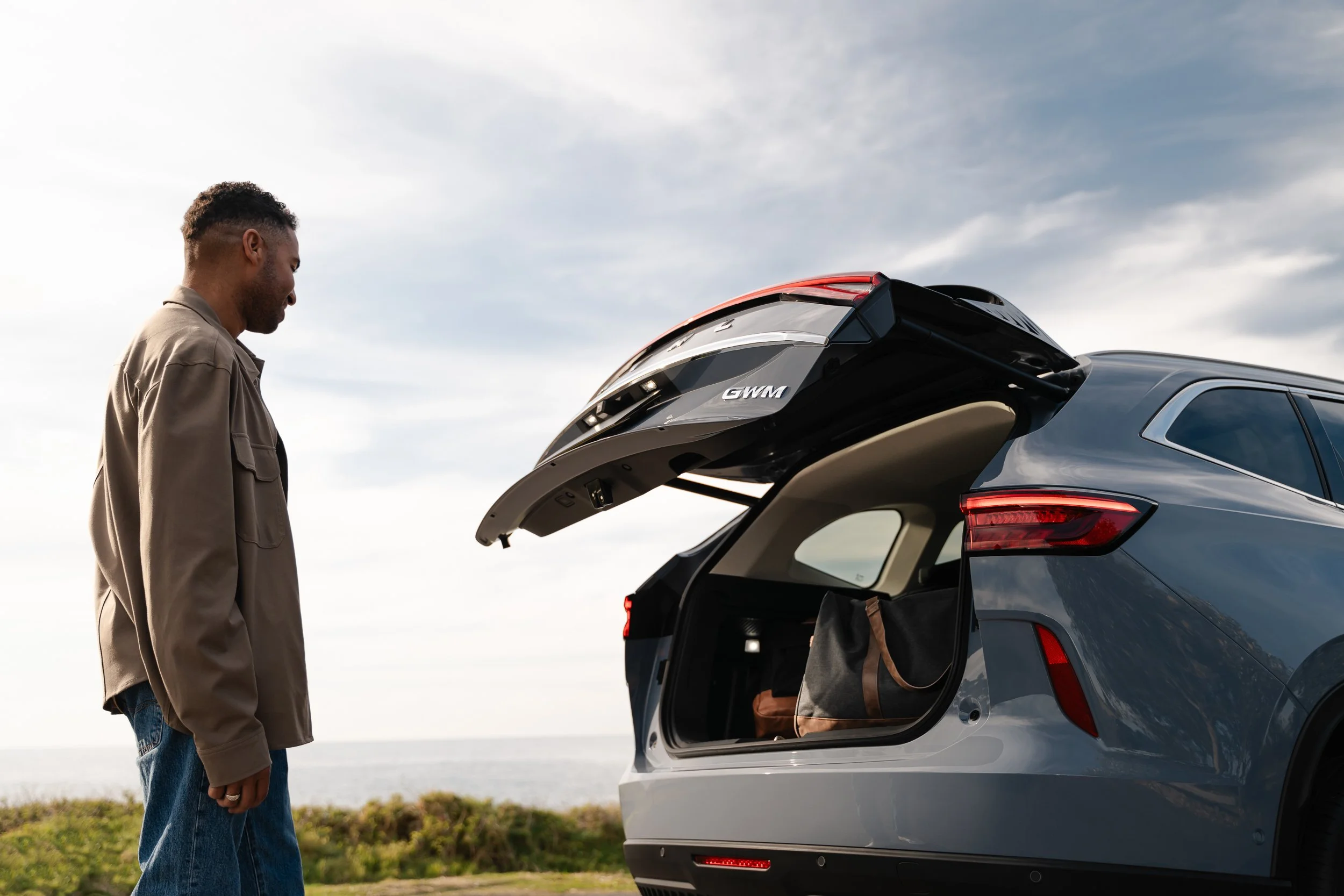 A man stands next to a silver SUV with its trunk open, looking inside. The setting appears to be outdoors near a body of water with a cloudy sky.