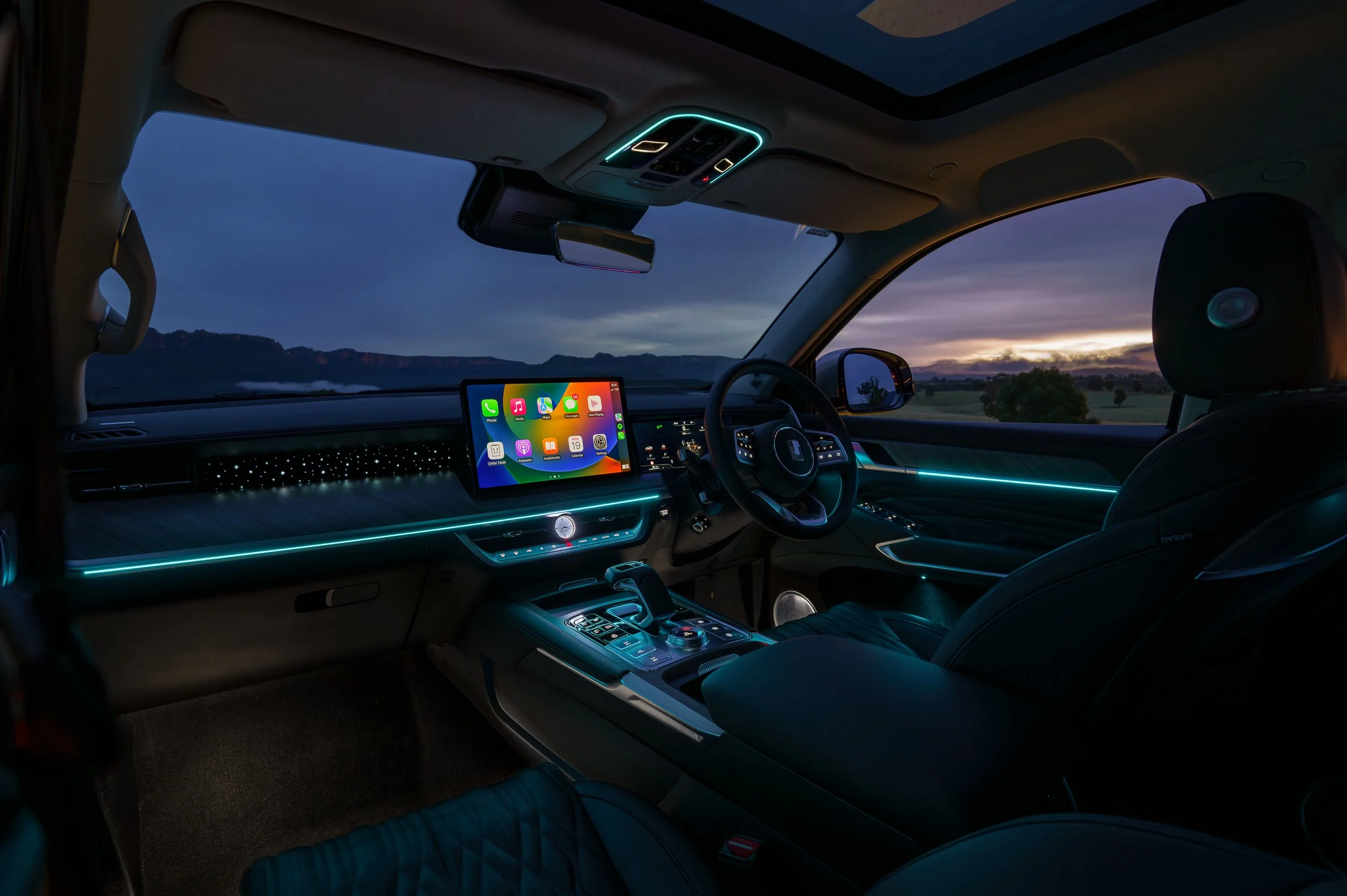 Interior of a modern car with ambient lighting, a digital dashboard, and a mounted screen displaying app icons, set against a scenic outdoor landscape at dusk visible through the windshield.
