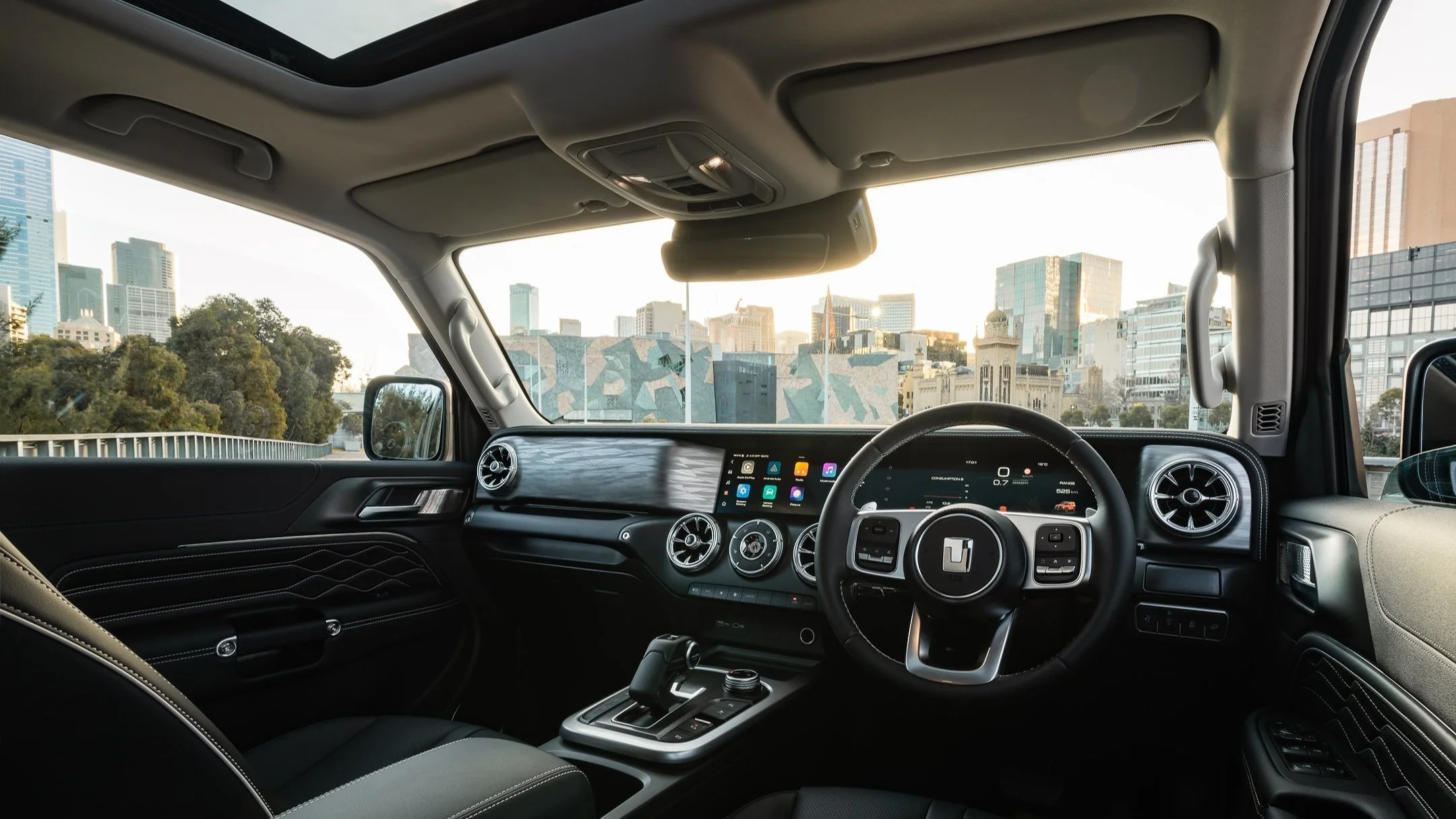 Interior view of a modern vehicle with a cityscape visible through the windshield, including tall buildings and a partly cloudy sky.