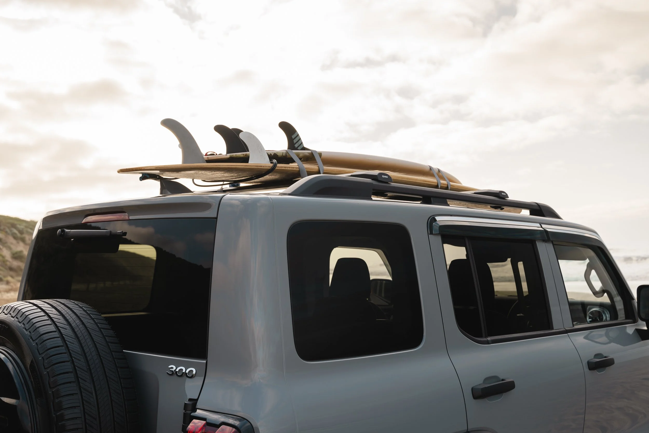 A silver SUV with a tire mounted on the back, parked outdoors with surfboards strapped on its roof rack, under a cloudy sky.
