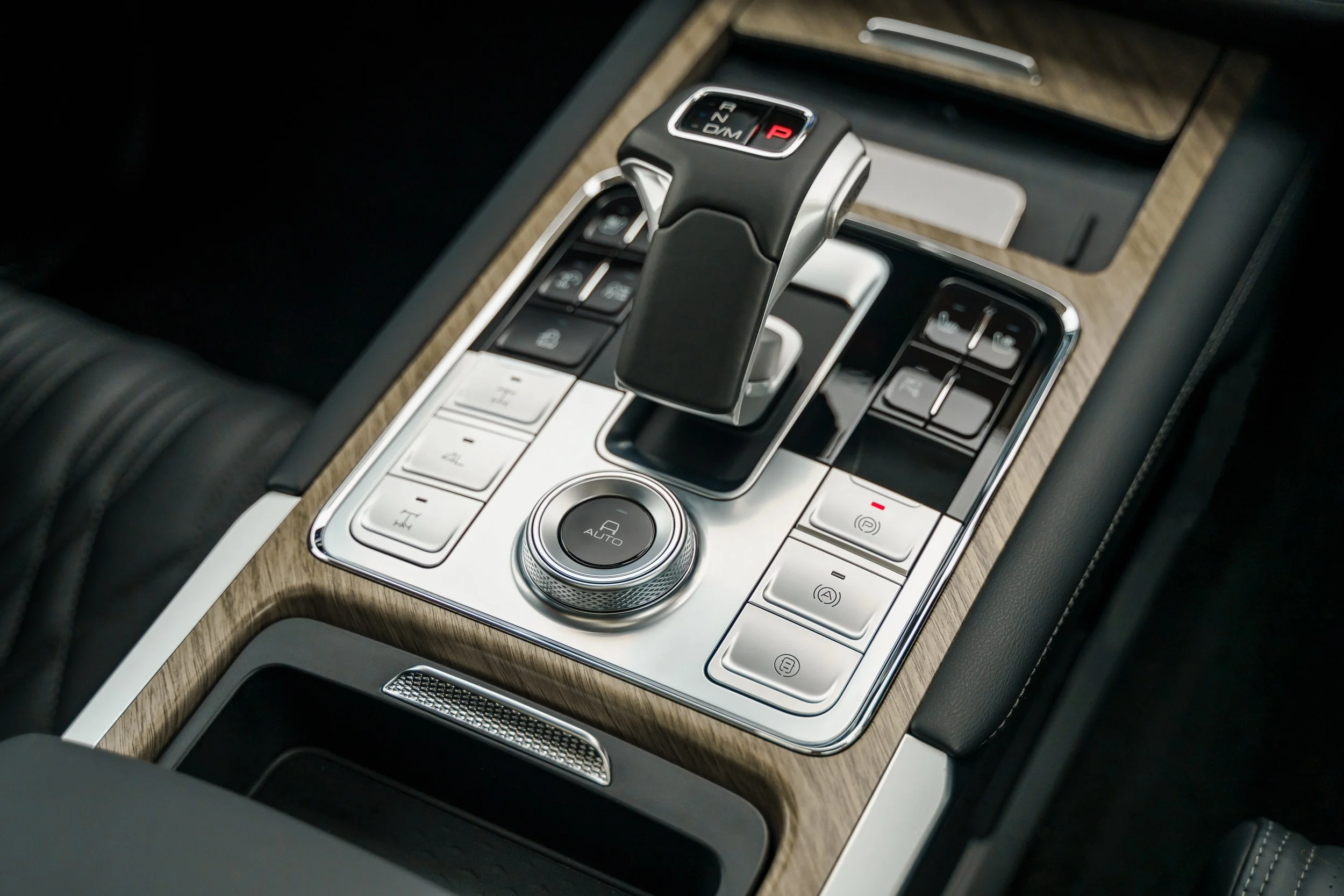 Close-up of a car's center console featuring a gear shift with P, R, N, D, and M modes, surrounded by control buttons, a rotary dial, and a wood trim surface.