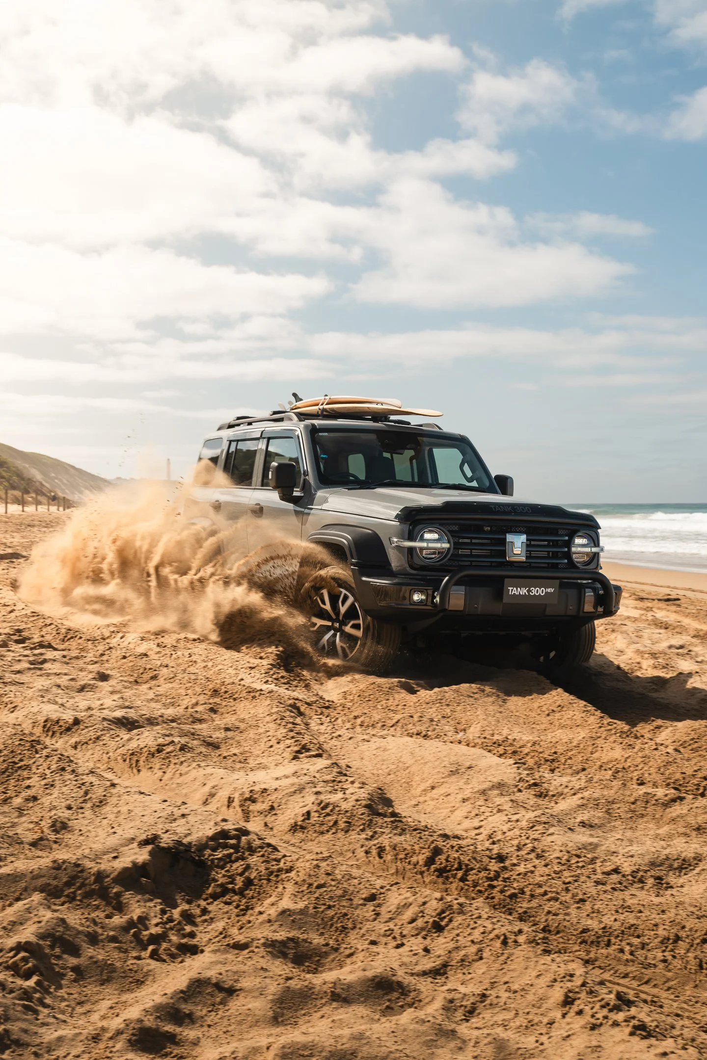 A black off-road vehicle driving on sandy beach, kicking up sand with surfboards mounted on roof, near ocean and cloudy sky.