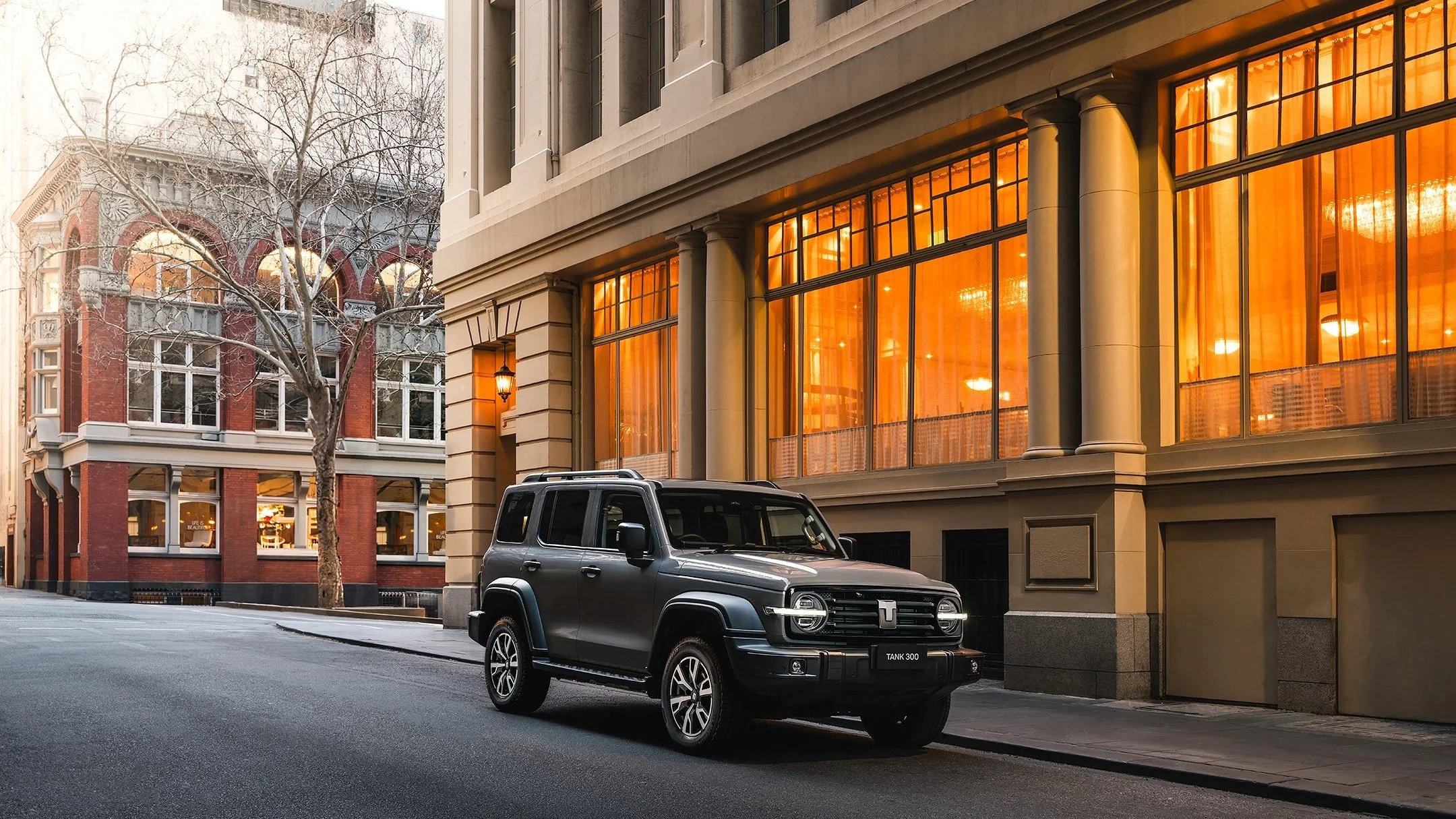 A black SUV parked on a city street in front of a modern building with large orange-lit windows. Another ornate, older building with white trim and leafless trees is visible in the background.