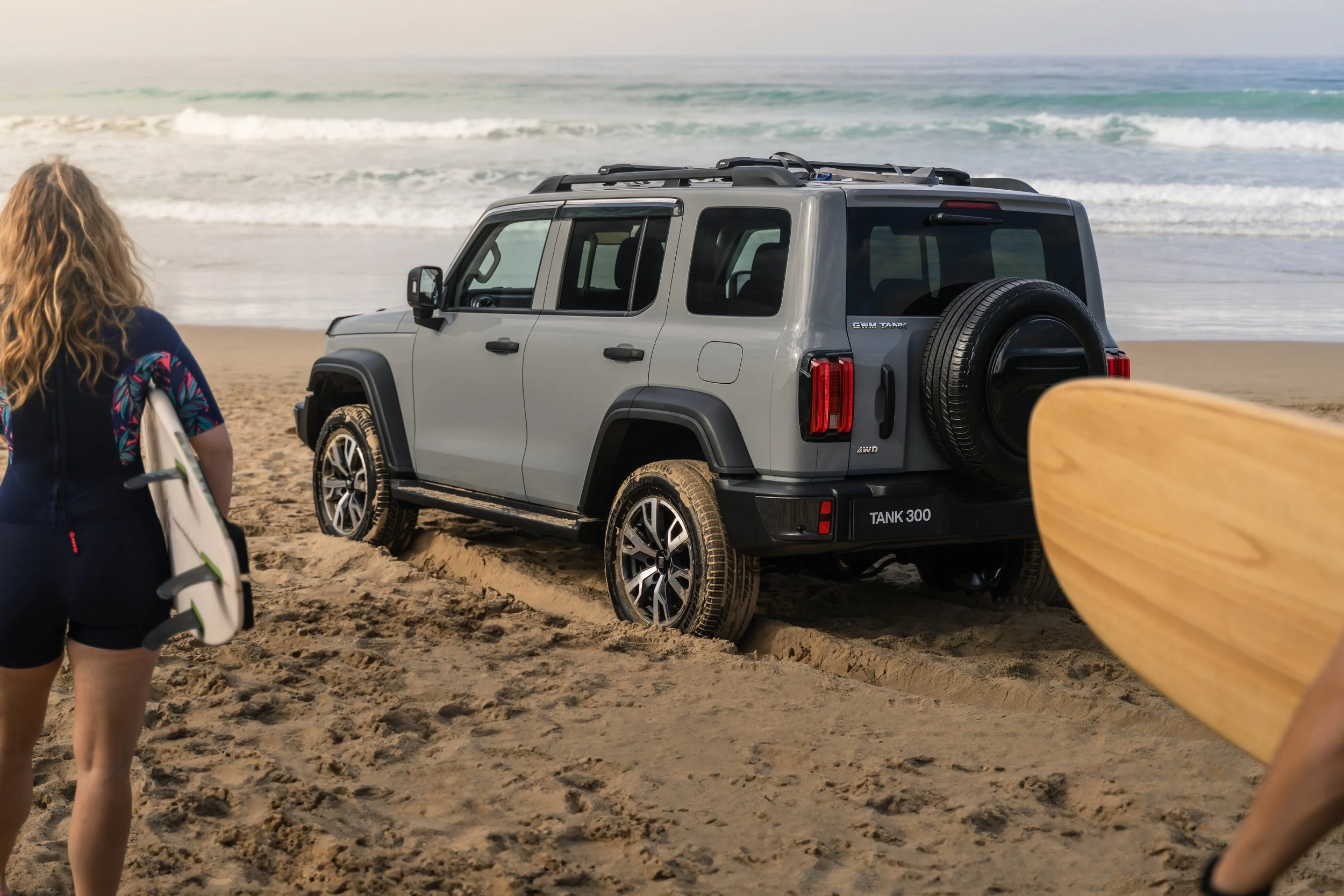 A person with long curly blonde hair holding a surfboard, standing on sandy beach near a silver SUV and ocean waves in the background.