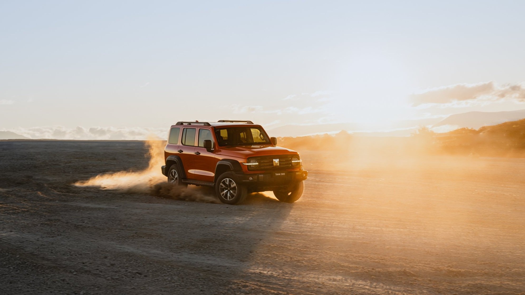 A red off-road SUV driving across a barren, dusty landscape at sunset.