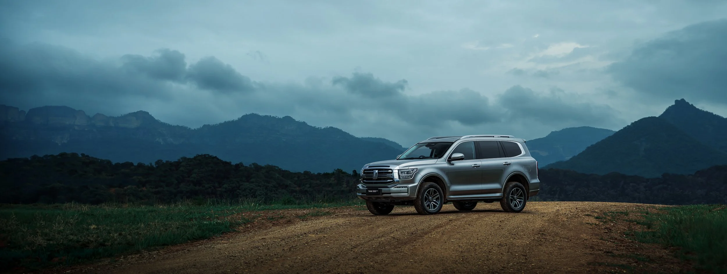 A silver SUV parked on a dirt road in front of a mountainous landscape with cloudy sky.