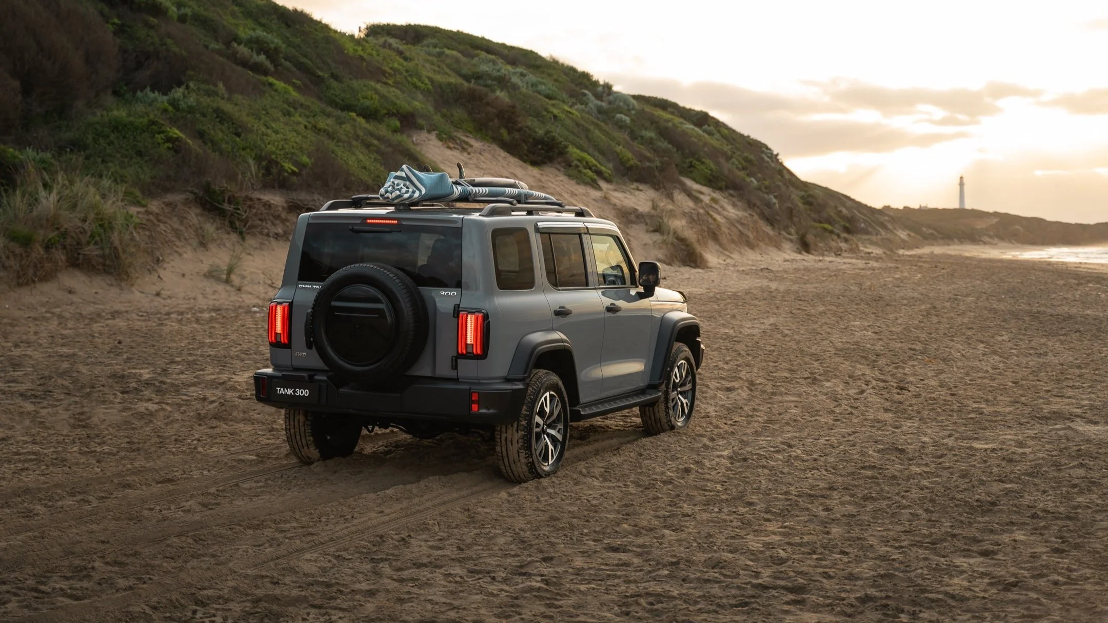 A gray off-road SUV parked on a sandy beach near green hills, with a surfboard on the roof, during sunset.