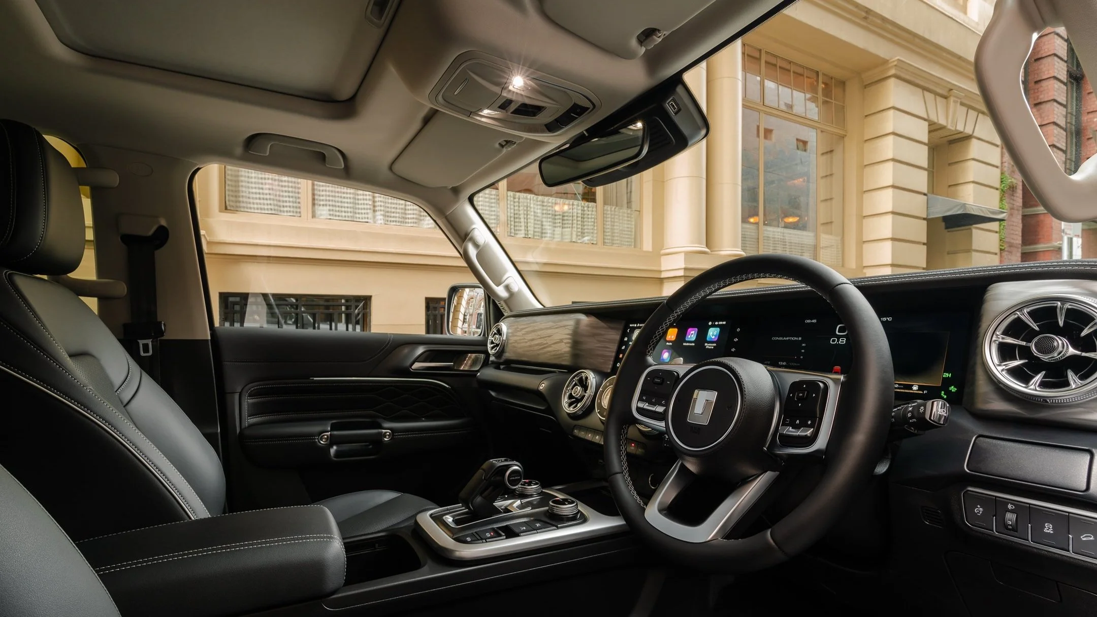 Interior of a modern vehicle showing the dashboard, steering wheel, and front seats, with large windows and classic building visible outside.