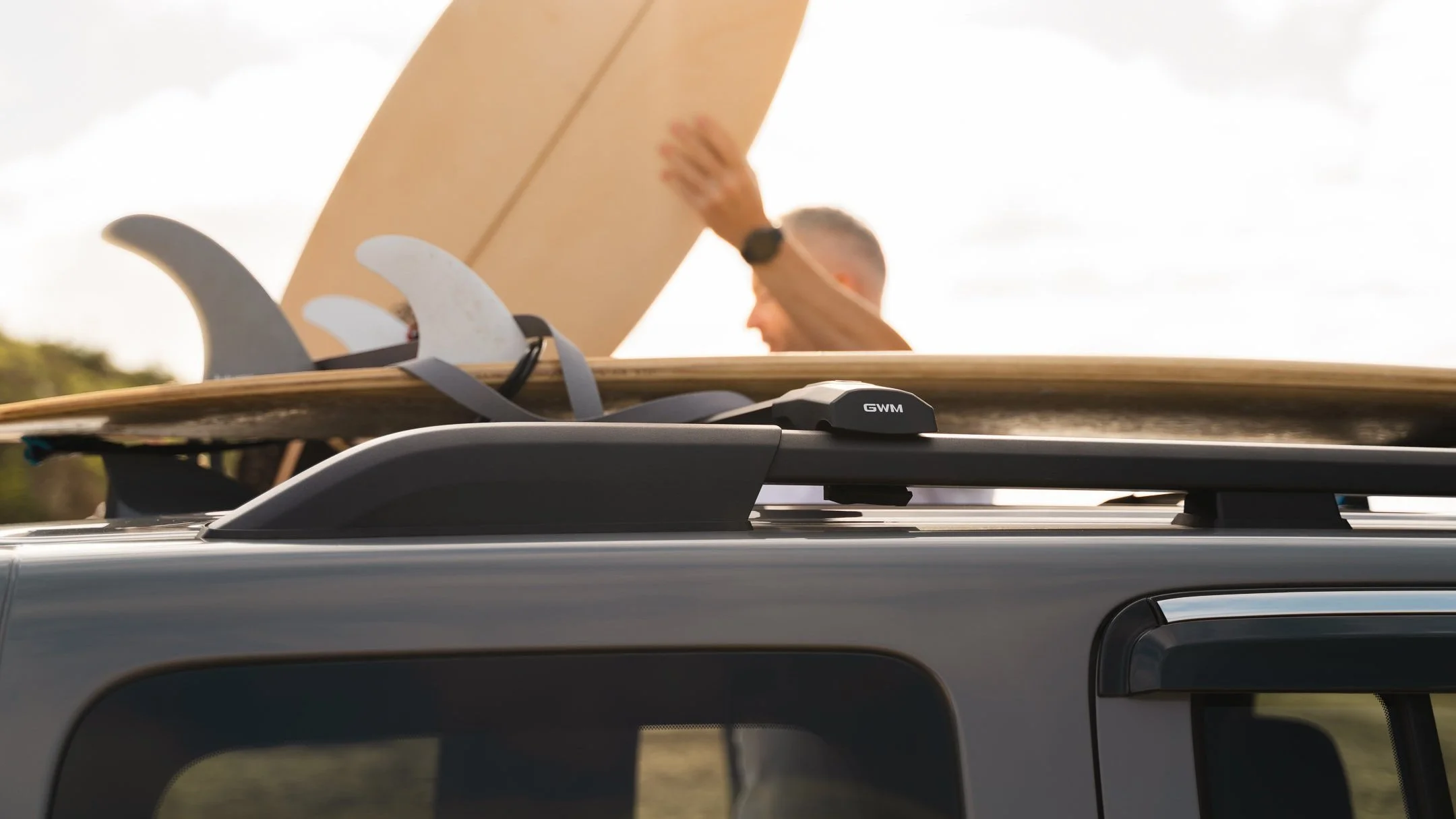 Close-up of a vehicle roof rack holding a surfboard and a snowboard, with a person in the background reaching towards the surfboard, during daytime.