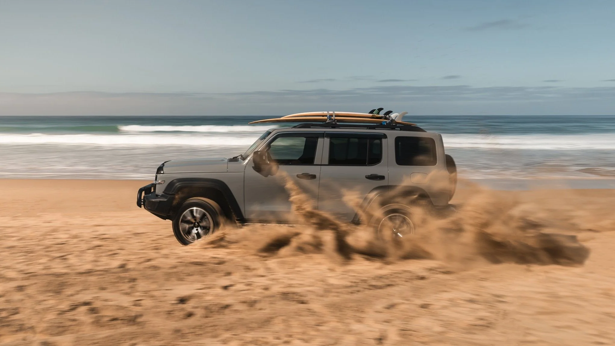 A black SUV driving on a sandy beach with surfboards attached to the roof, kicking up sand as it moves near the ocean.
