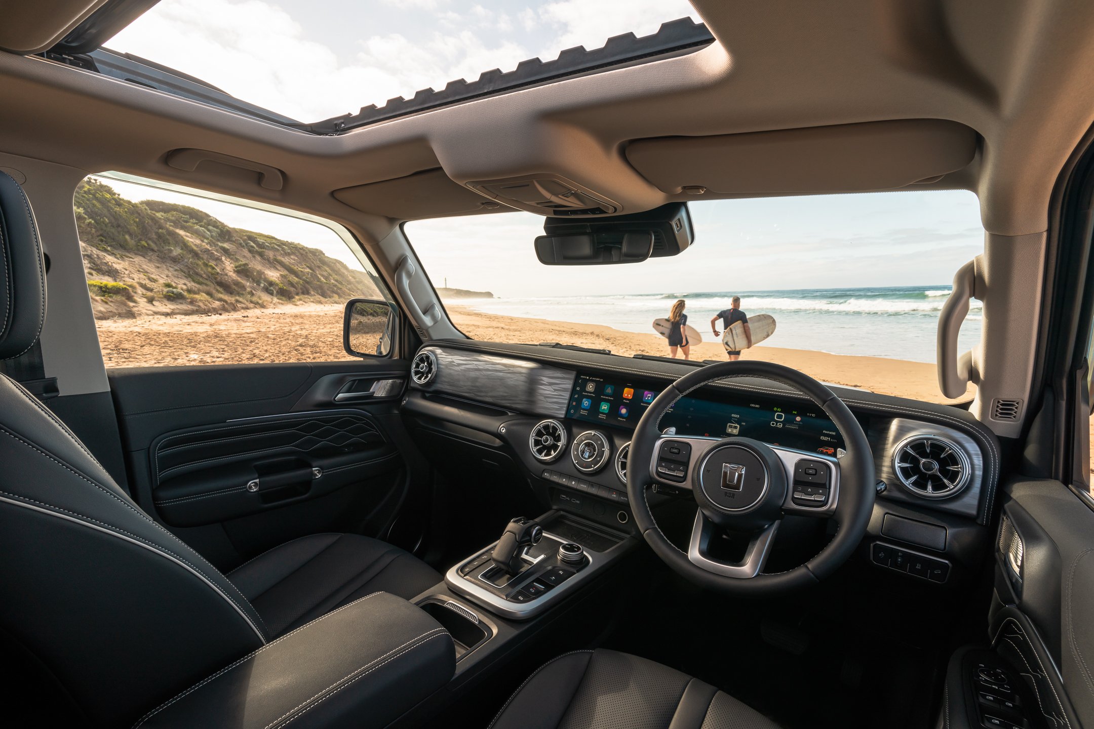 View from inside a vehicle looking out through the windshield at a sandy beach with two people carrying surfboards and walking towards the ocean.