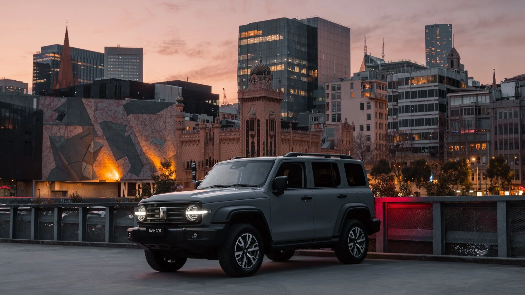 A gray SUV parked on a city street during dusk with a skyline of modern and historic buildings in the background, including a church tower and a clock tower.
