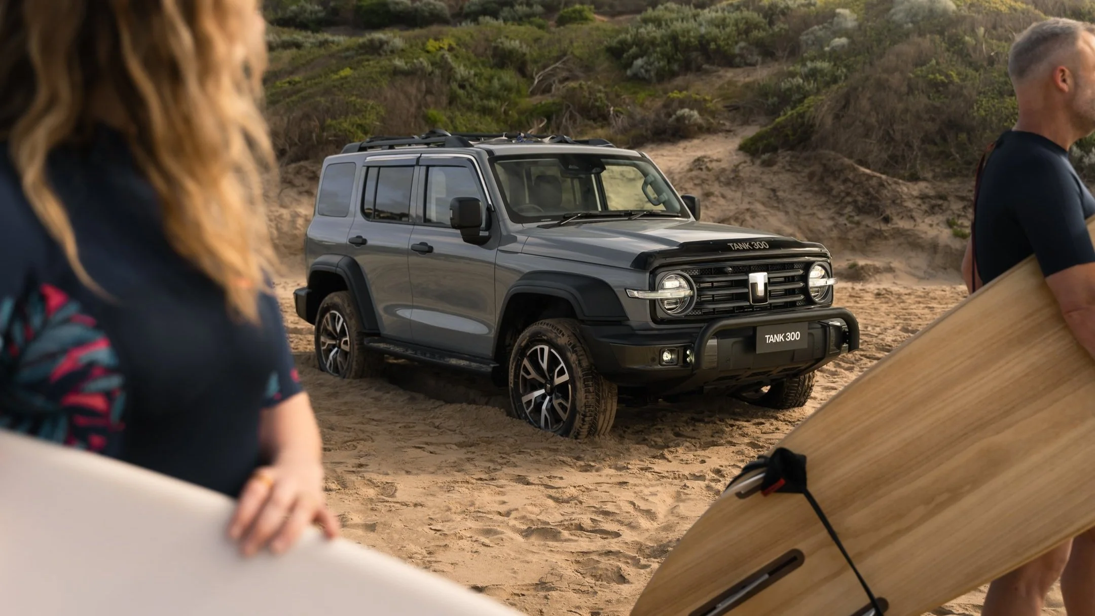 A gray off-road vehicle on sandy terrain with bushes and hills in the background, surrounded by people, one holding a surfboard.