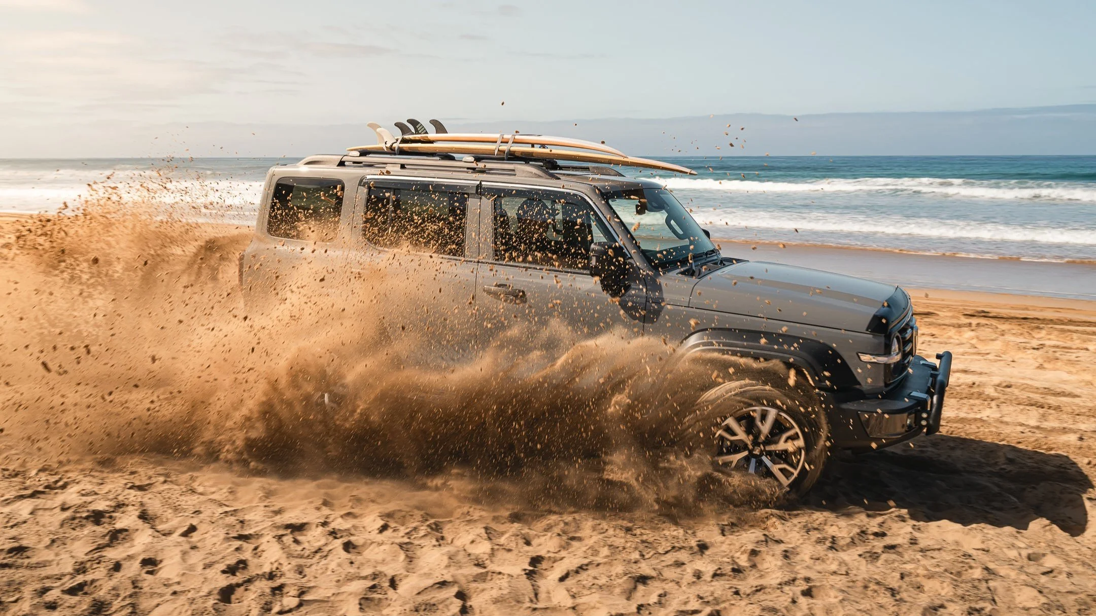 A black SUV driving on the beach, kicking up sand with surfboards on the roof, near the ocean with waves and a cloudy sky.
